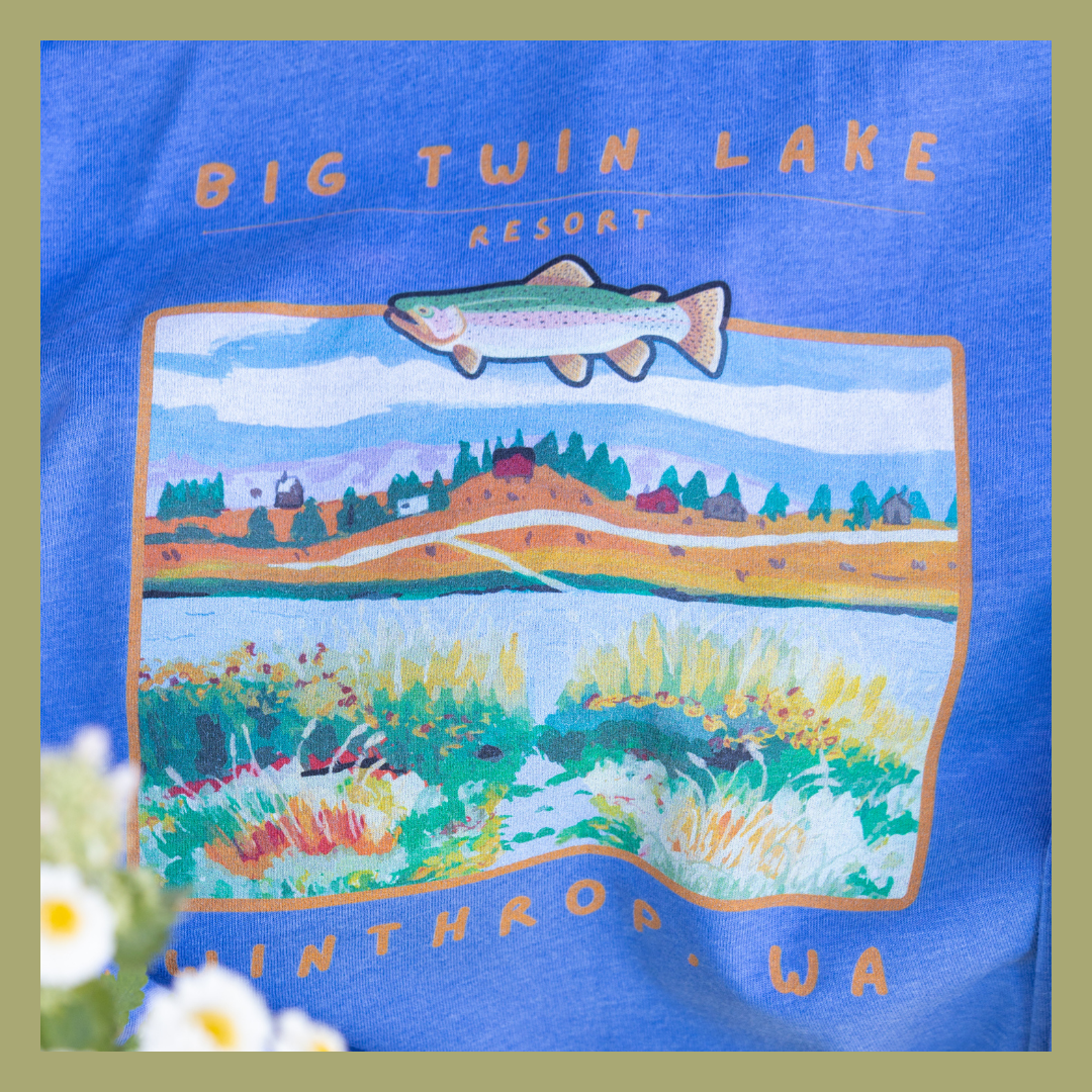 A colorful graphic design on a blue shirt featuring a lake landscape with mountains, trees, and flowers, and a fish illustration above, with the text 'Big Twin Lake Resort' and 'Whitrow WA'.
