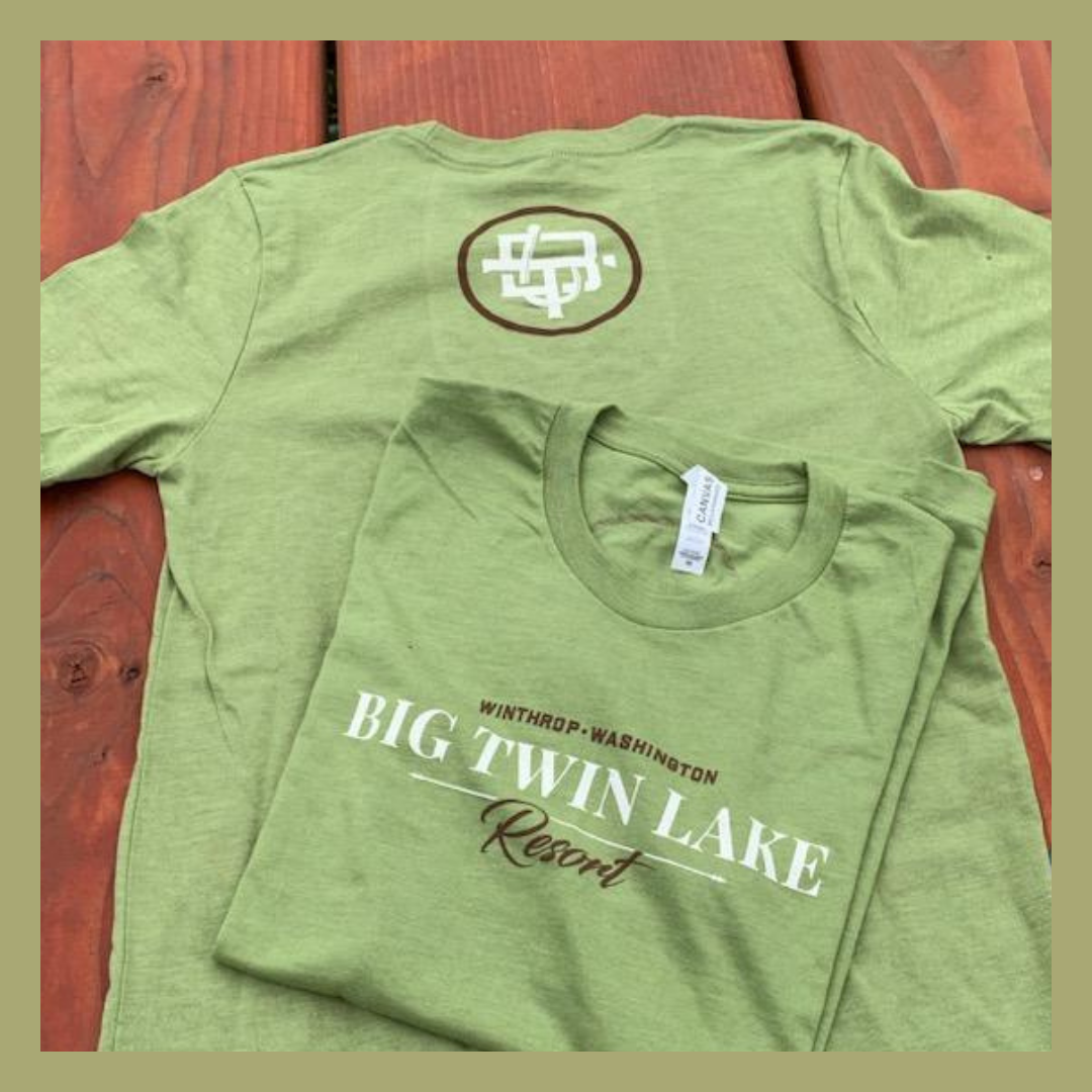 Green t-shirts, one folded and one laid out, with the text "BIG TWIN LAKE Resort" and "WINTHROP WASHINGTON" printed on the front, resting on a wooden surface.
