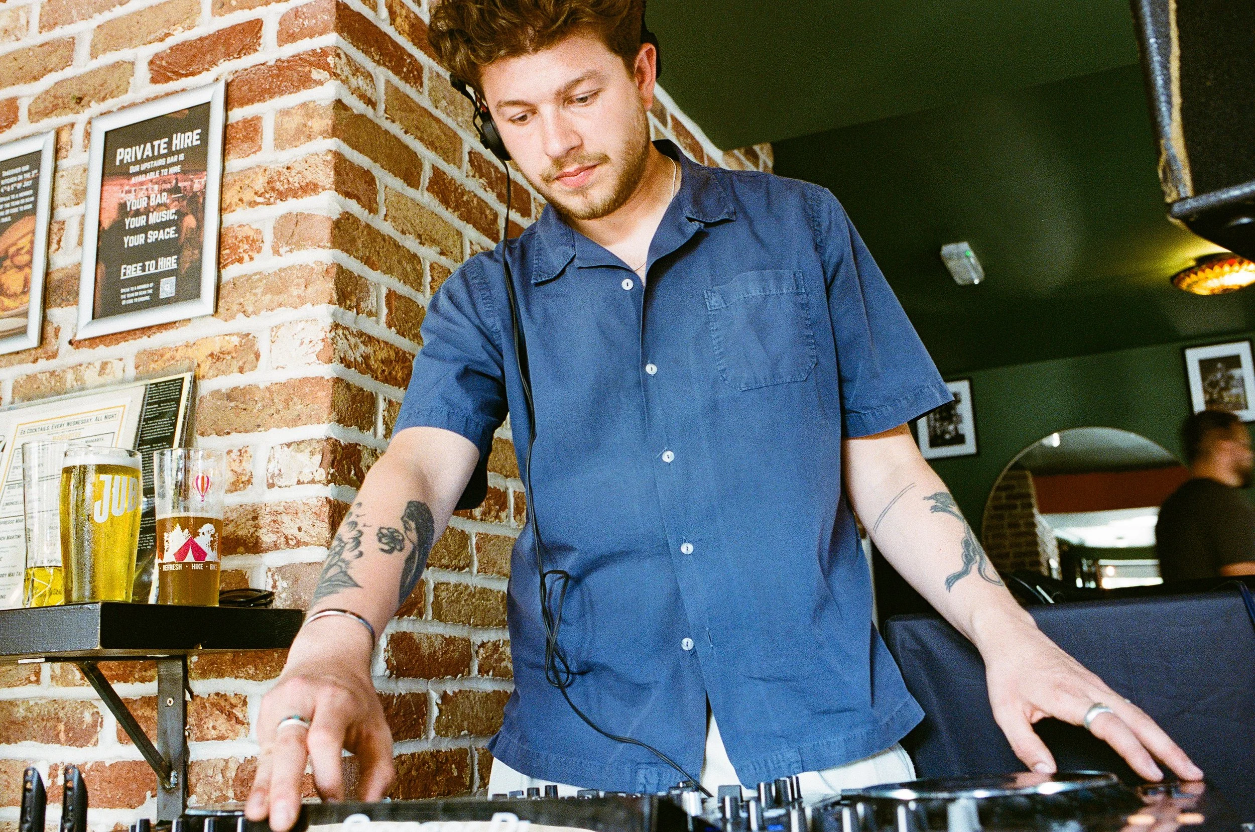 A young man with curly hair and tattoos on his arms is DJing at a bar or restaurant, wearing headphones and adjusting equipment.