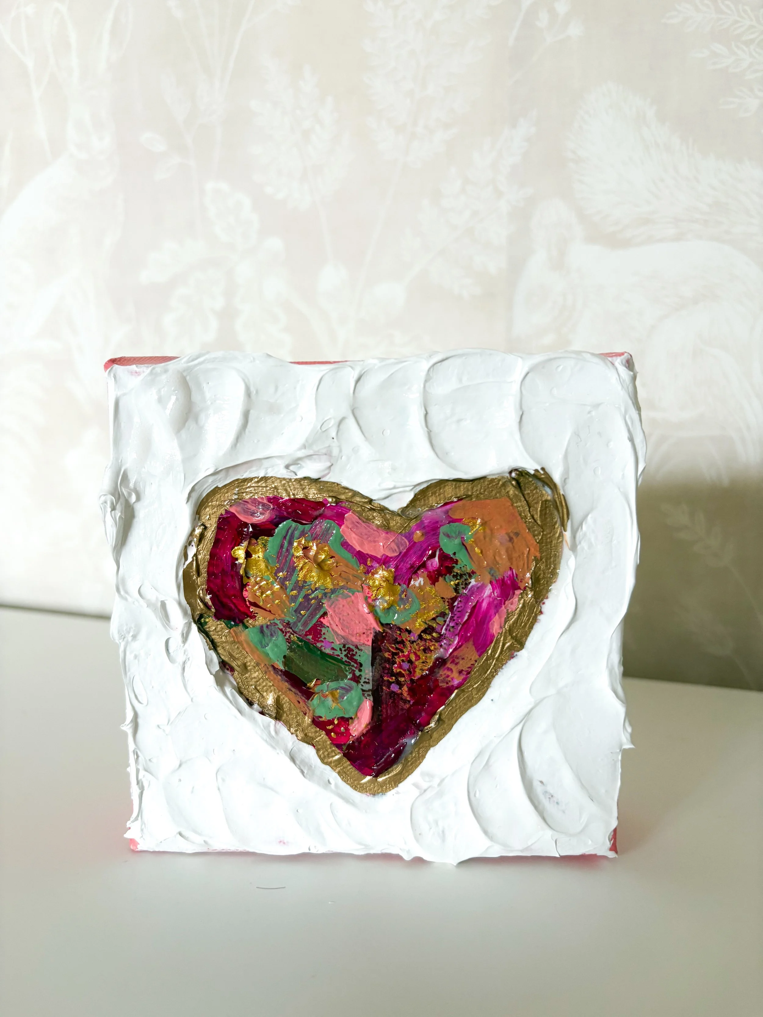 A small canvas with a textured white background featuring a colorful, abstract heart outlined in gold and filled with vibrant brush strokes of pink, green, and red.