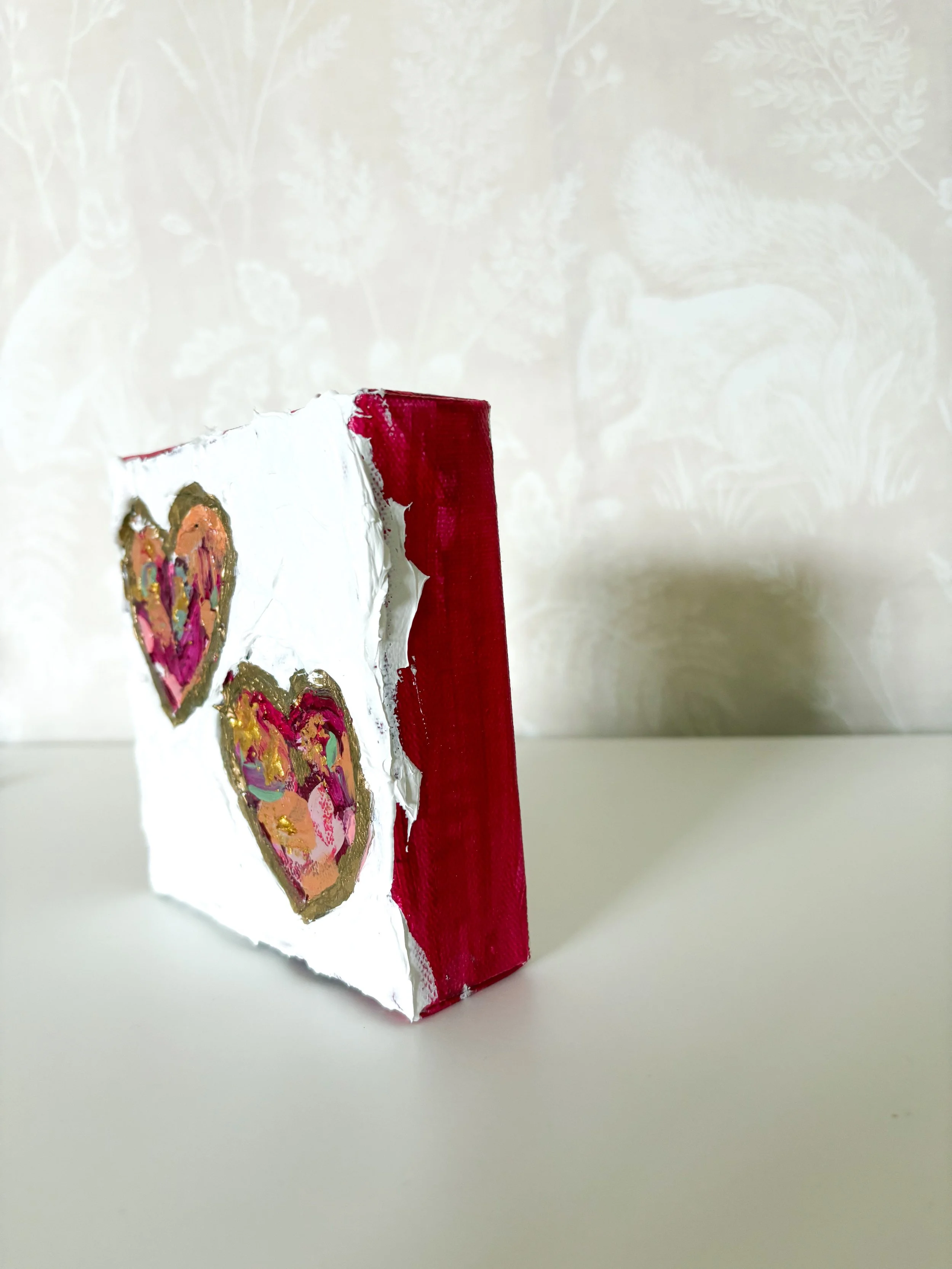 A small, rectangular canvas painting of two colorful hearts with gold accents on a white background, placed on a white surface in front of a patterned beige wall.