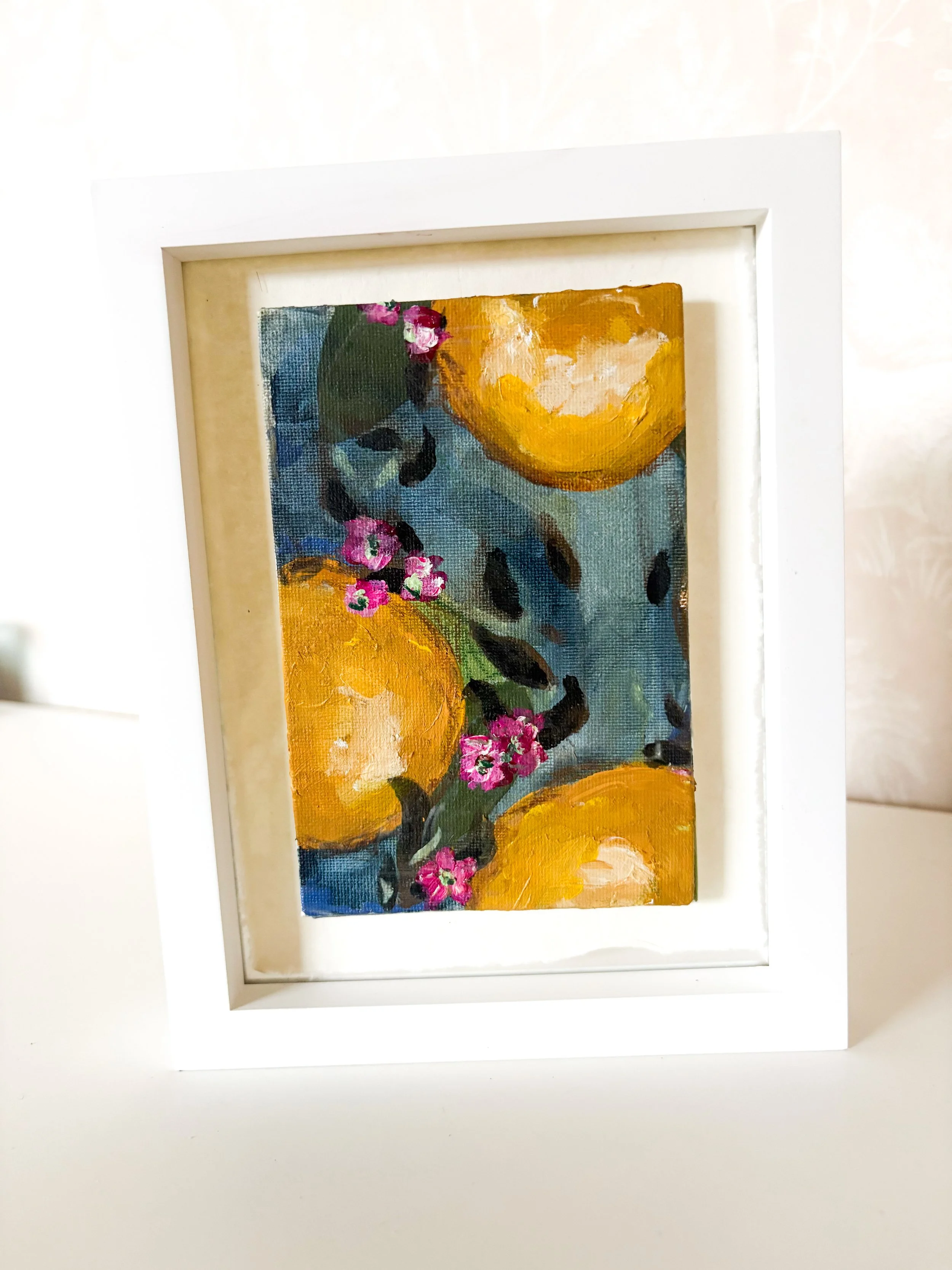 Small framed painting of three large yellow oranges, green leaves, and pink flowers on a blue background.