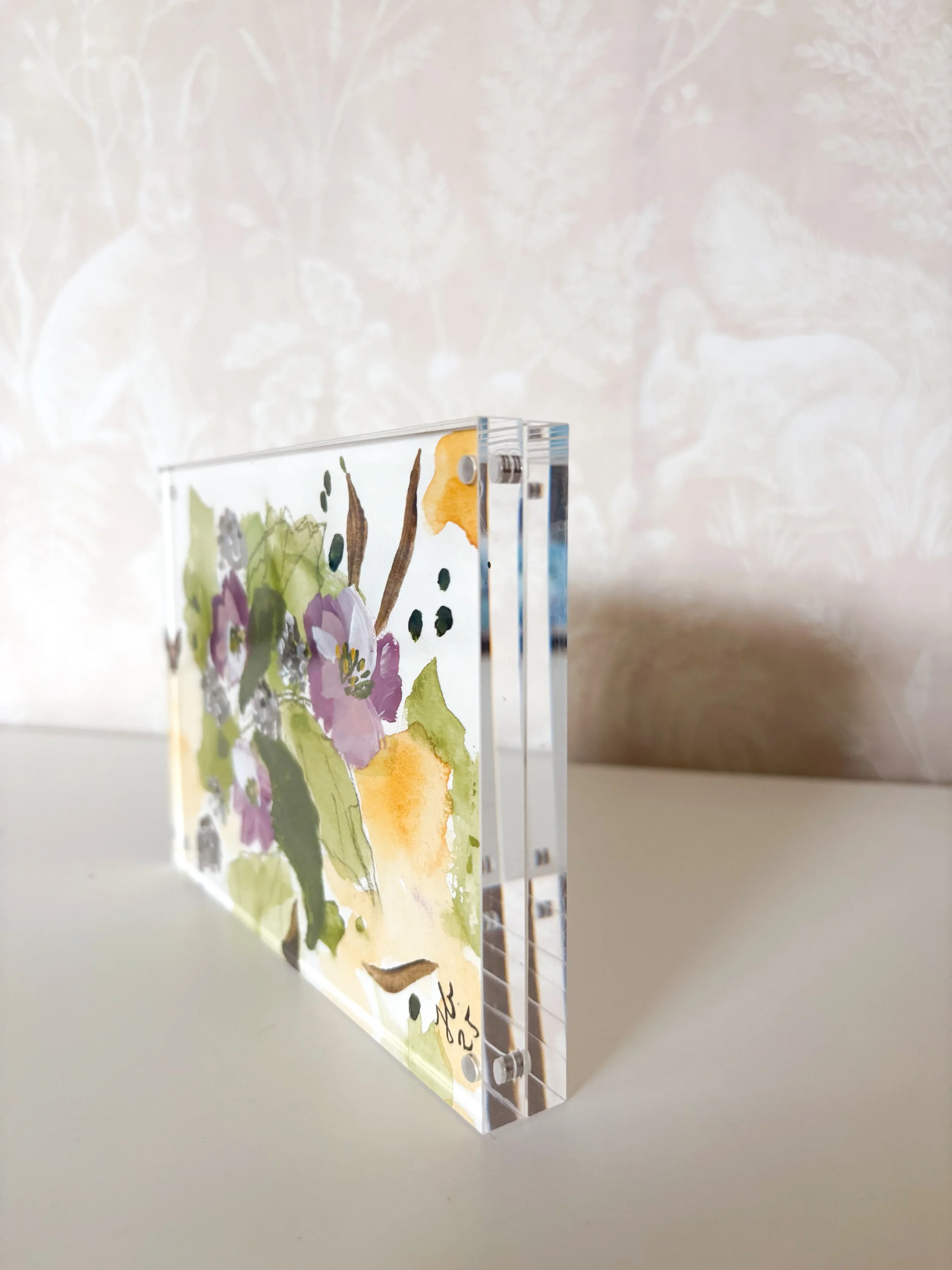 A rectangular acrylic art piece with a watercolor floral design featuring purple, orange, yellow, and green colors, standing upright on a white surface with a beige patterned wall in the background.