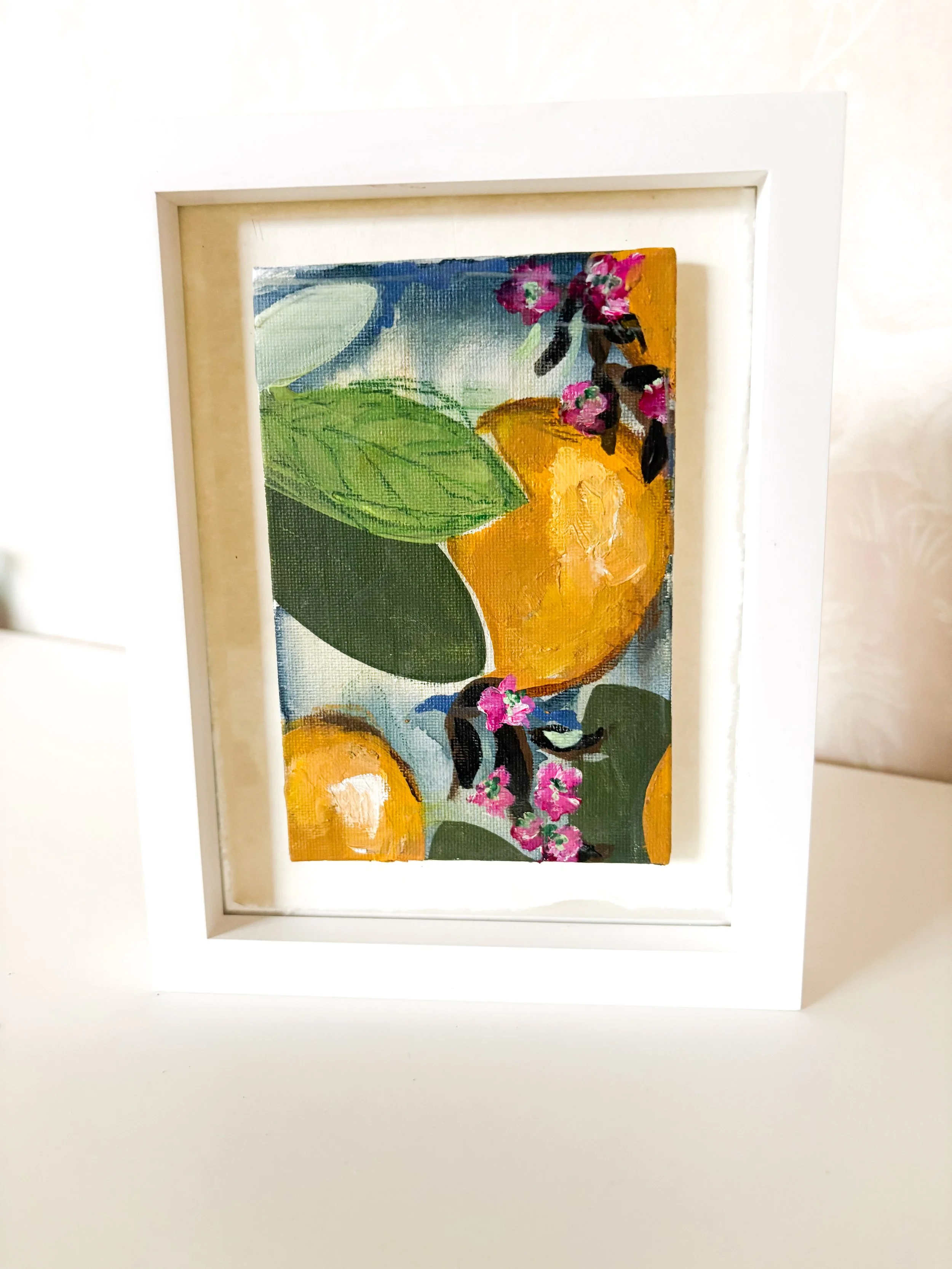 A framed painting of oranges, pink flowers, and green leaves on a blue background.