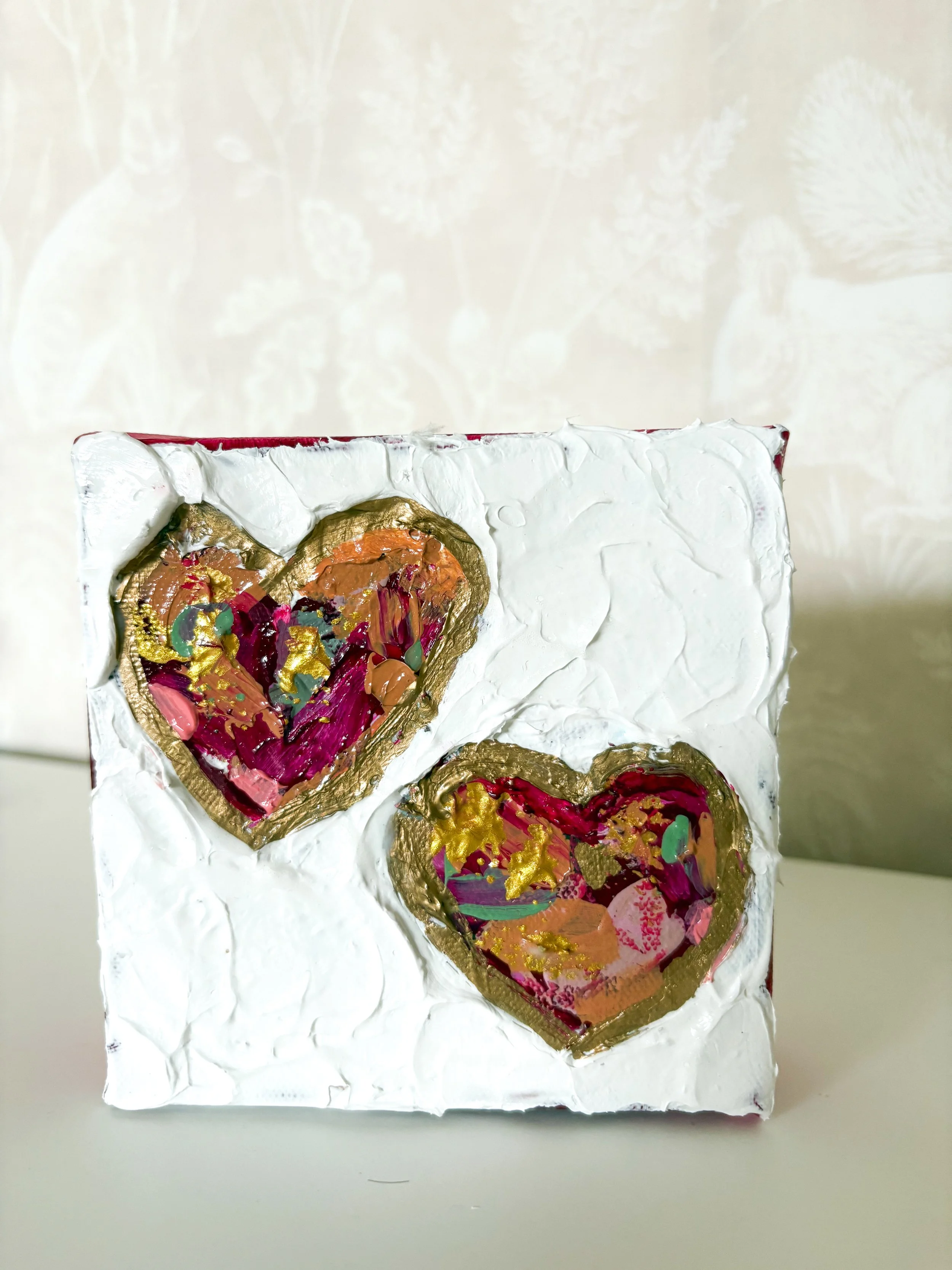 Decorative 3D canvas with two abstract hearts outlined in gold, filled with colorful paint splatters and gold accents against a textured white background.