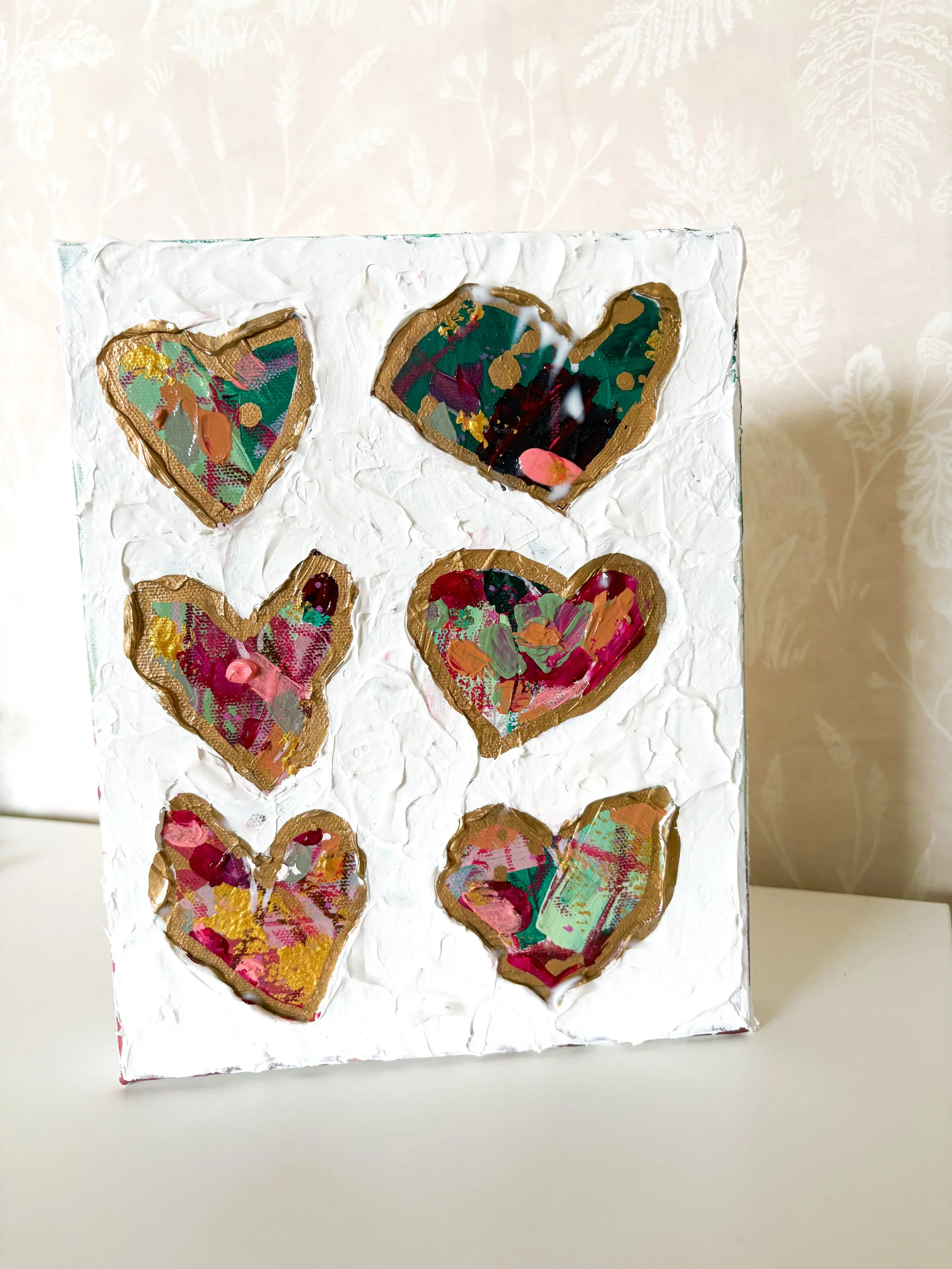 An art canvas with six painted hearts outlined in gold, set against a textured white background.