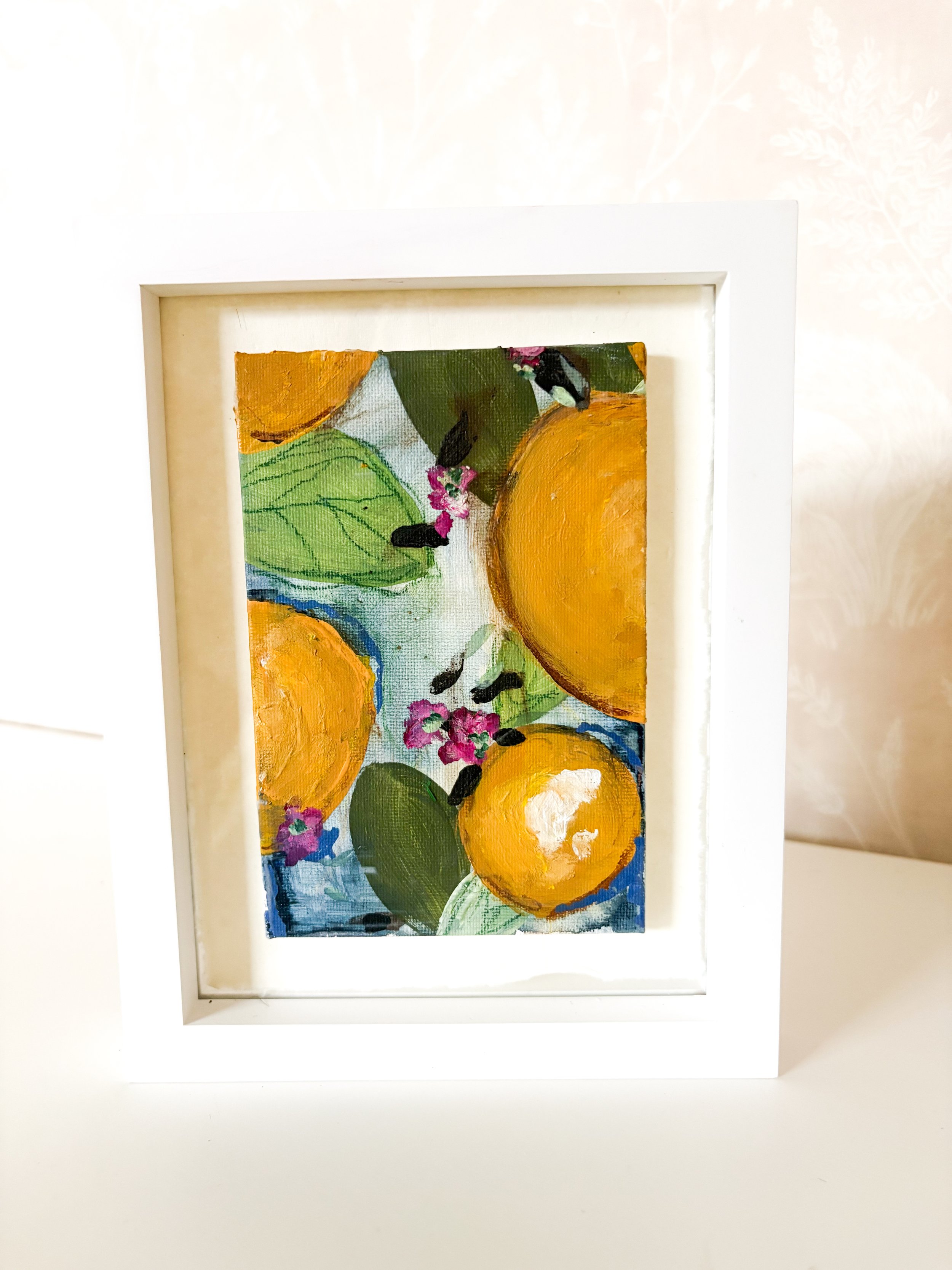 Painting of oranges, green leaves, and small pink flowers in a white frame.