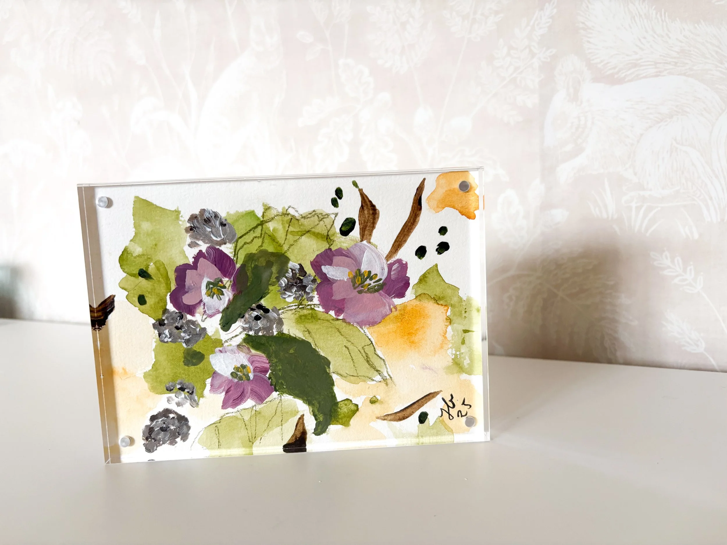 A watercolor painting of flowers and leaves on a clear acrylic panel, with a white surface and a white wallpaper background with a subtle botanical pattern.