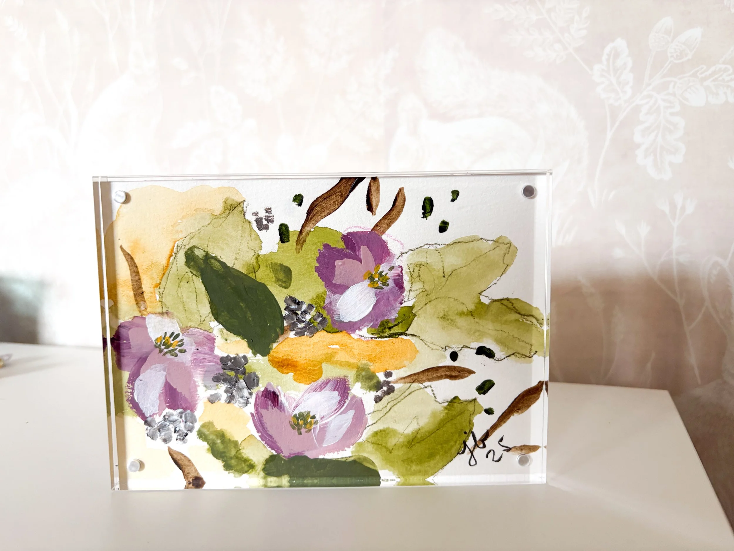 A transparent acrylic panel features a watercolor painting of purple flowers, green leaves, and brown stems, placed on a white surface with a light-colored, patterned wall in the background.