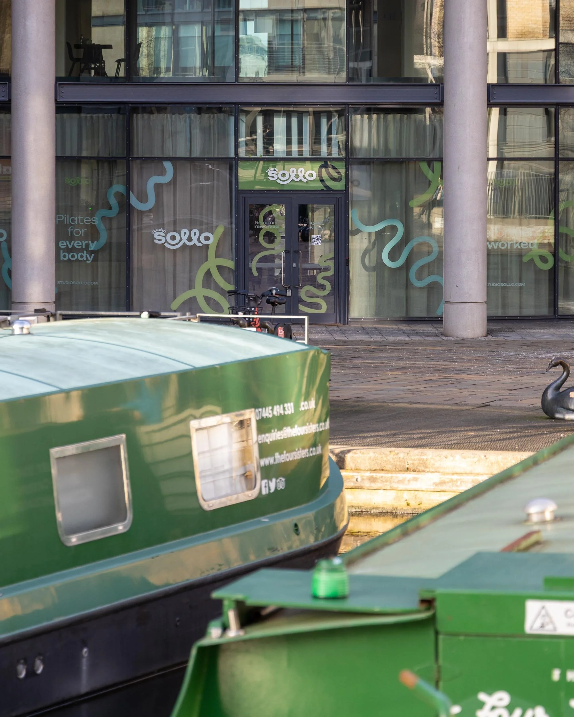Exterior view of Studio Sollo fitness studio with large glass windows and colorful snake graphics, building entrance with glass door, and bicycles parked outside.