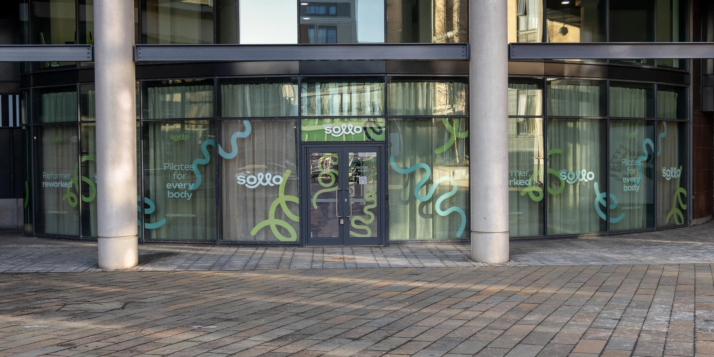 The storefront of Studio Sollo features glass windows decorated with the studio's logo and colorful abstract squiggles. The windows also display the phrases "Reformer, reworked," "Pilates for everybody," and the studio's website, studiosollo.com.