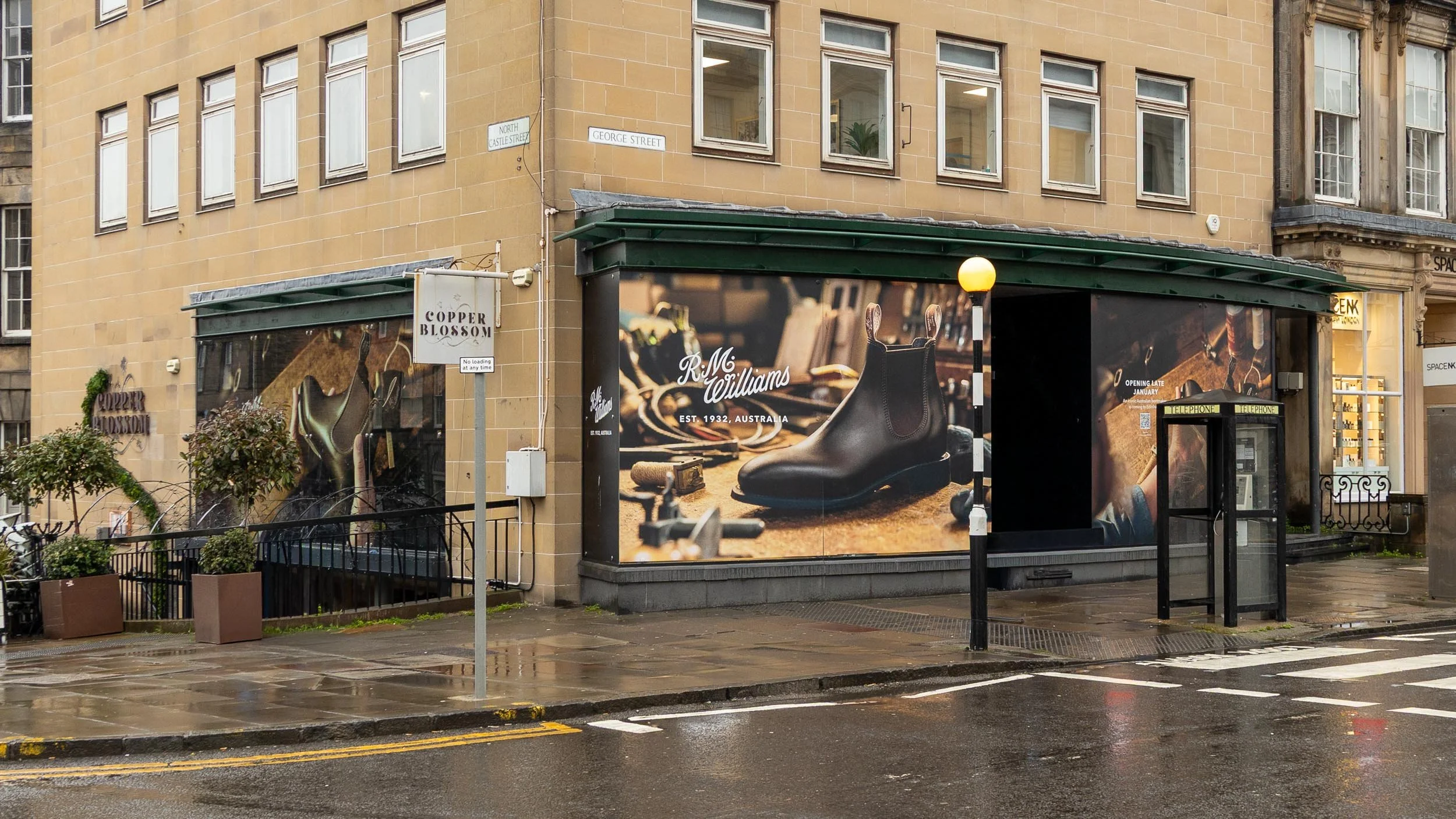 Street corner with a storefront advertising R.M. Williams boots, featuring a large display of a black boot with a background of boot-making tools, a yellow streetlamp, a telephone booth, planters with small trees, and wet pavement.