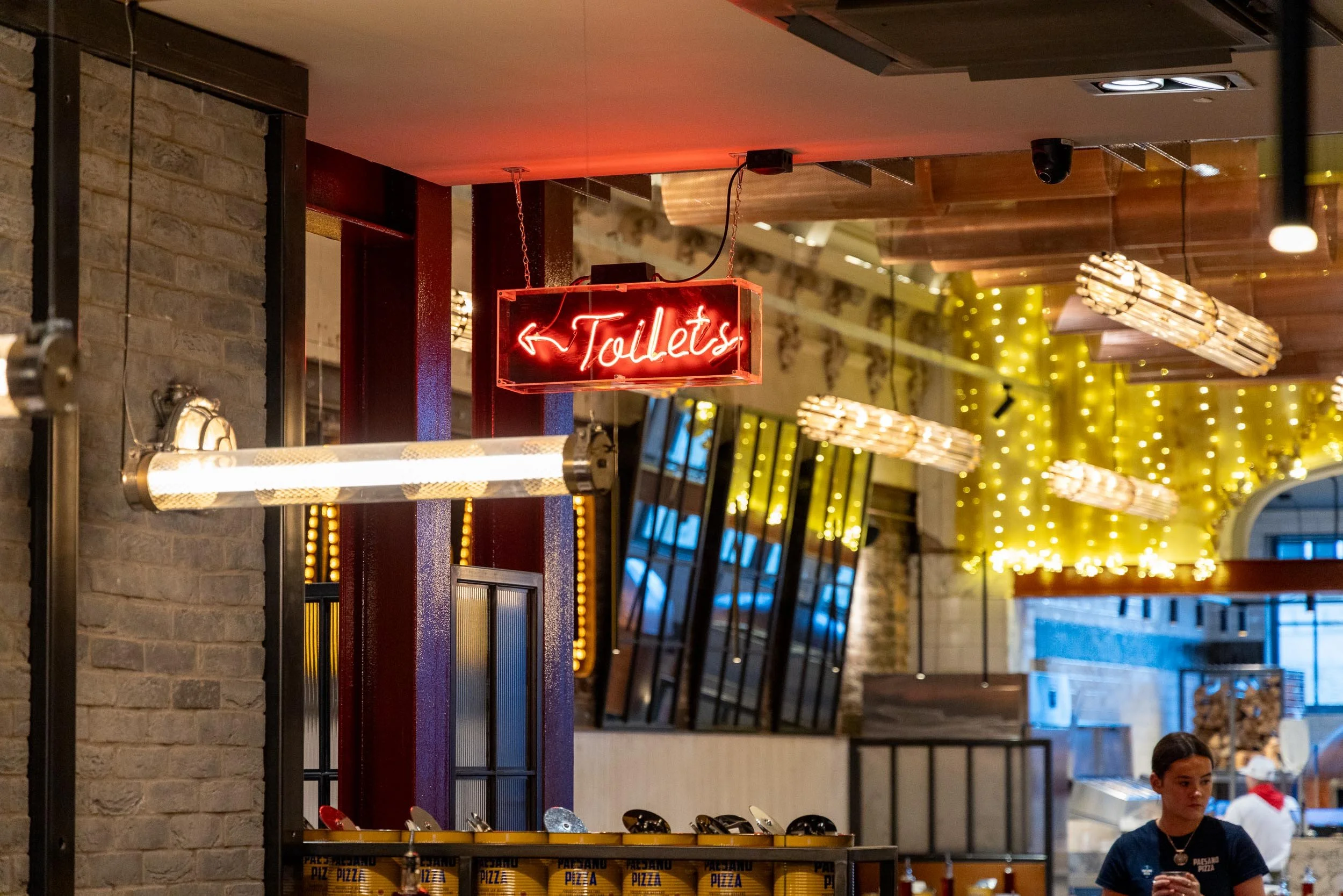 Indoor restaurant or cafe with warm lighting, a neon sign pointing to toilets, and a woman standing near a pizza counter.