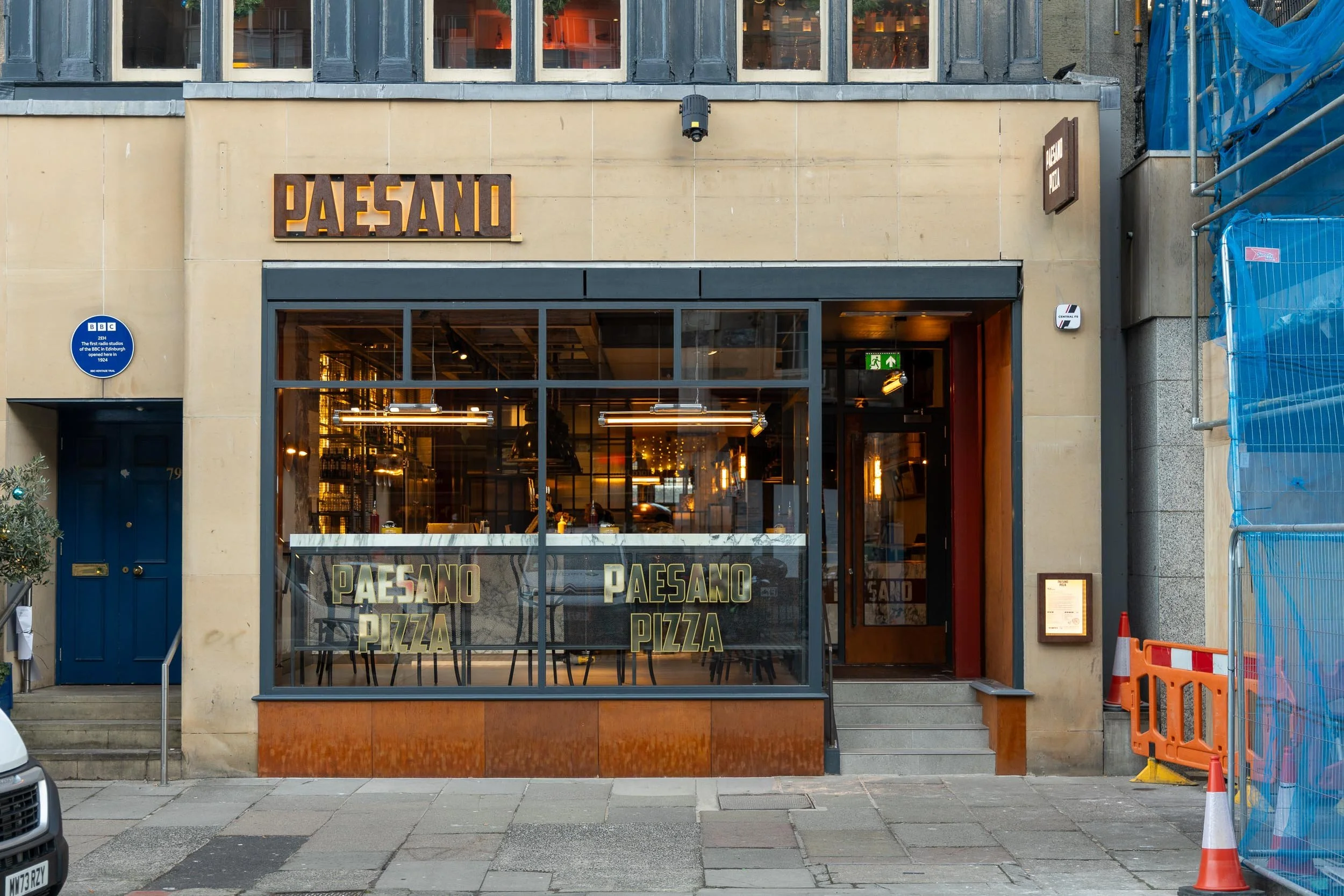 The exterior of a restaurant called Paesano, with large windows displaying the interior and the name Paesano Pizza on the glass. The restaurant is on an urban street with steps leading to the entrance, and there are some construction barriers on the 