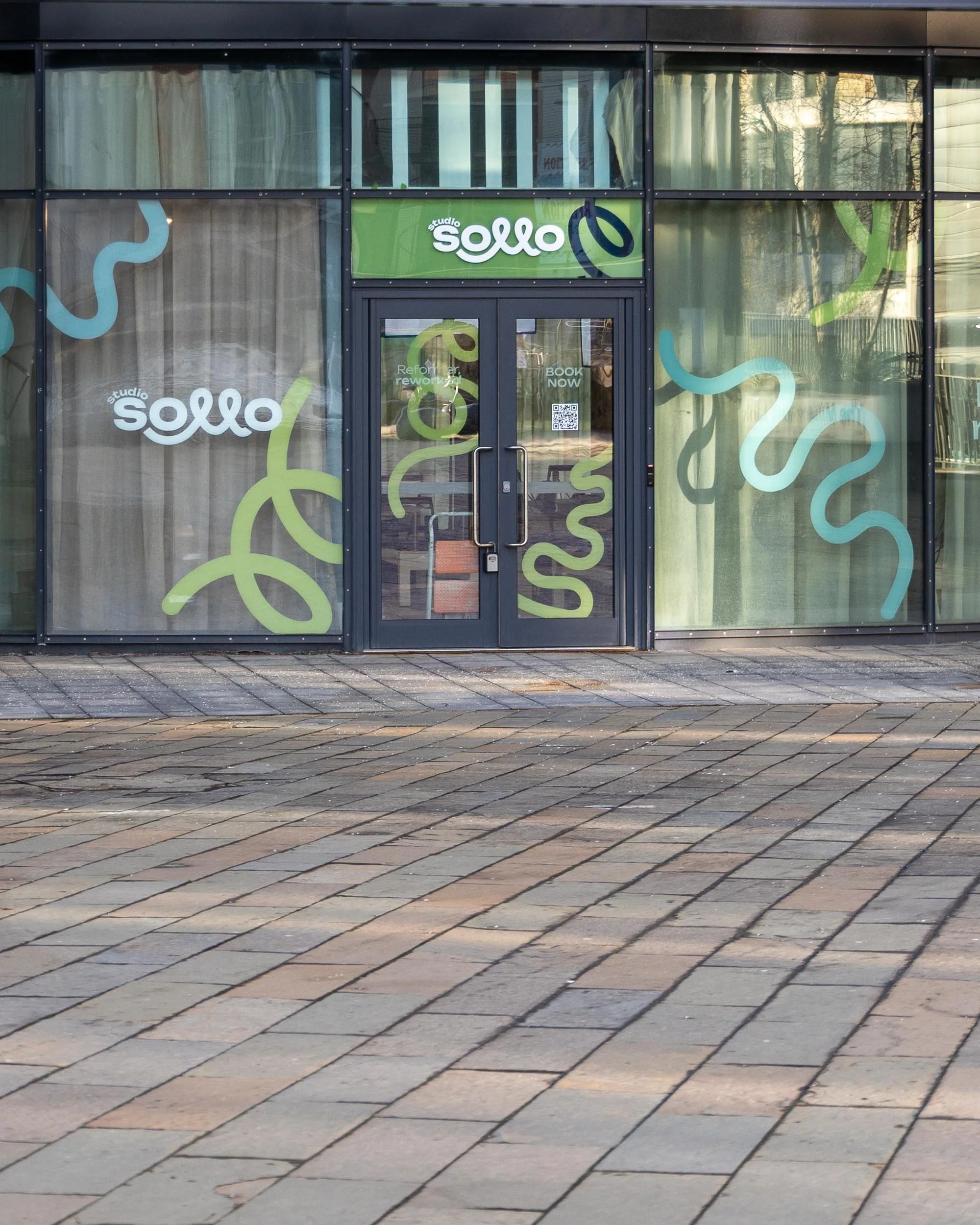 Front view of a modern glass storefront with the sign 'studio sollo' above the door, decorated with abstract squiggly line graphics in green, blue, and white.