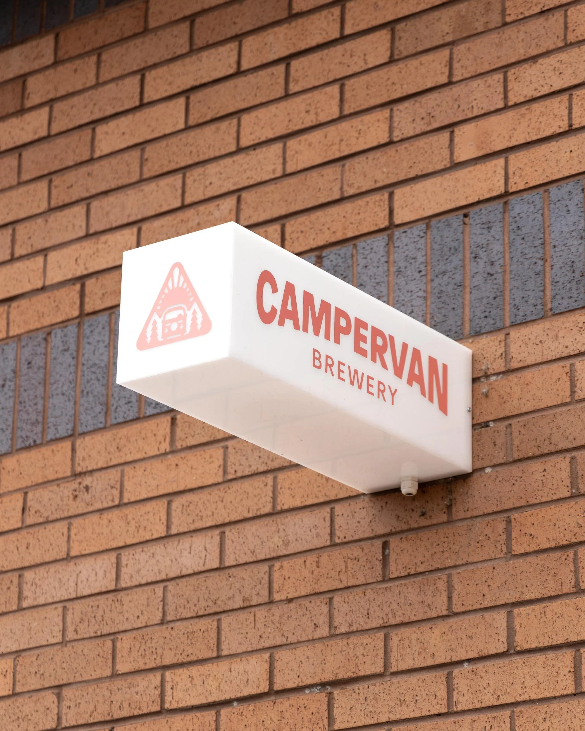 A white rectangular sign with red letters reading 'CAMPERVAN BREWERY' mounted on a brick wall. The sign has a logo of a camper van with trees inside a triangle.