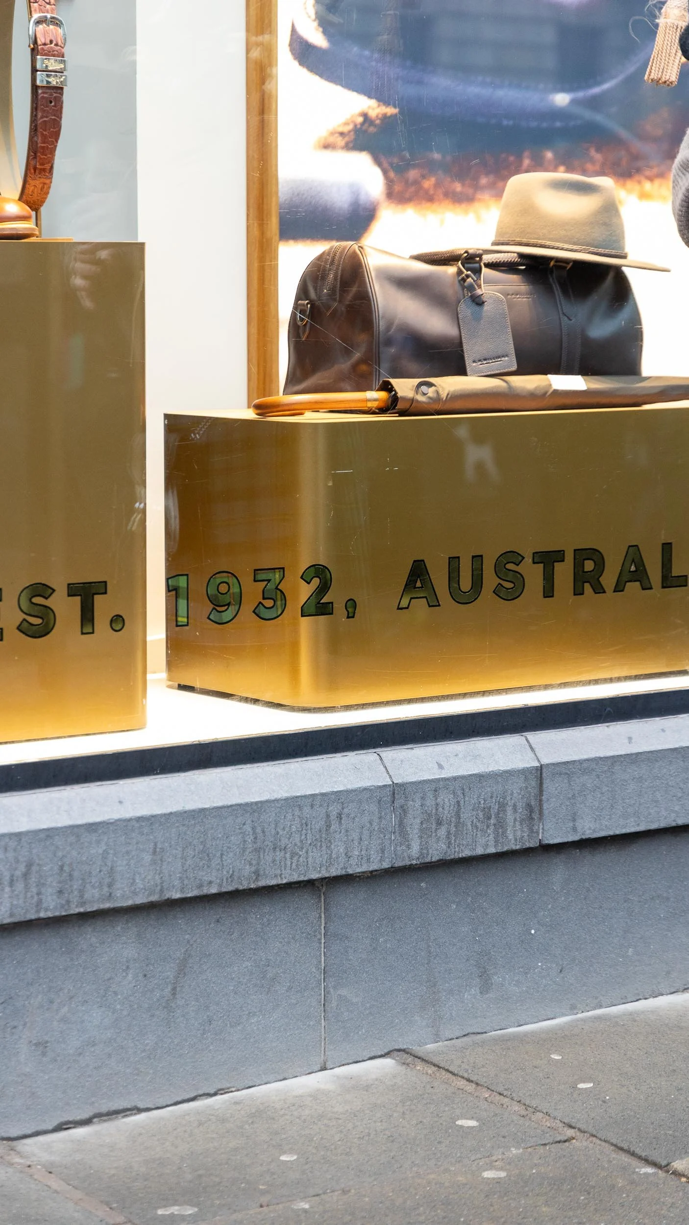 Store display window with hand applied glass gilded lettering 'EST. 1932, AUSTRALIA' and product display behind, including a black duffel bag, a beige hat, and a black umbrella.