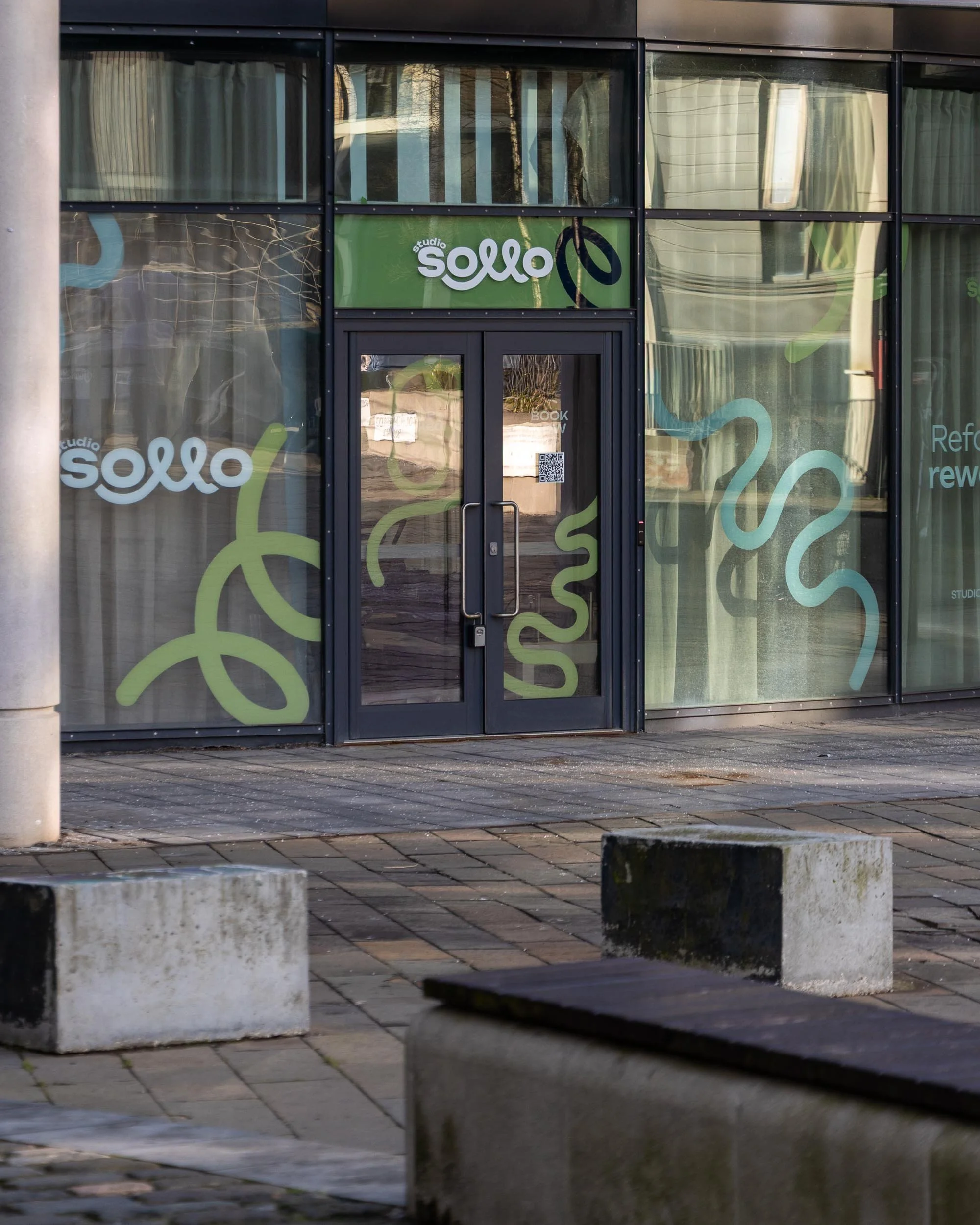 Front view of a glass building entrance with the signage 'Studio Sollo' and decorative green and blue lines on the windows.