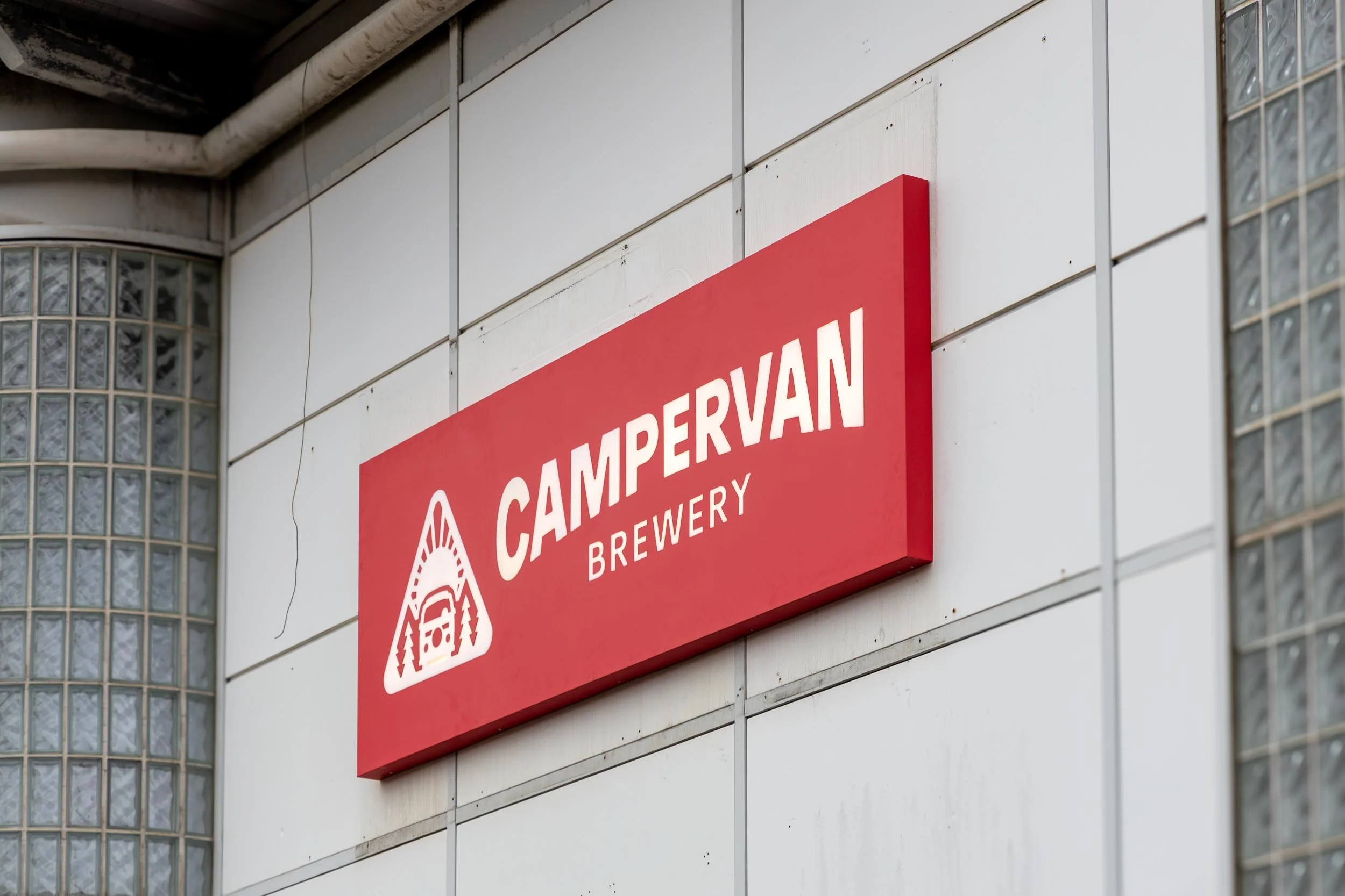 Red sign with white text reading 'CAMPERVAN BREWERY' and a logo of a camper van within a triangular shape, mounted on the exterior wall of a building.