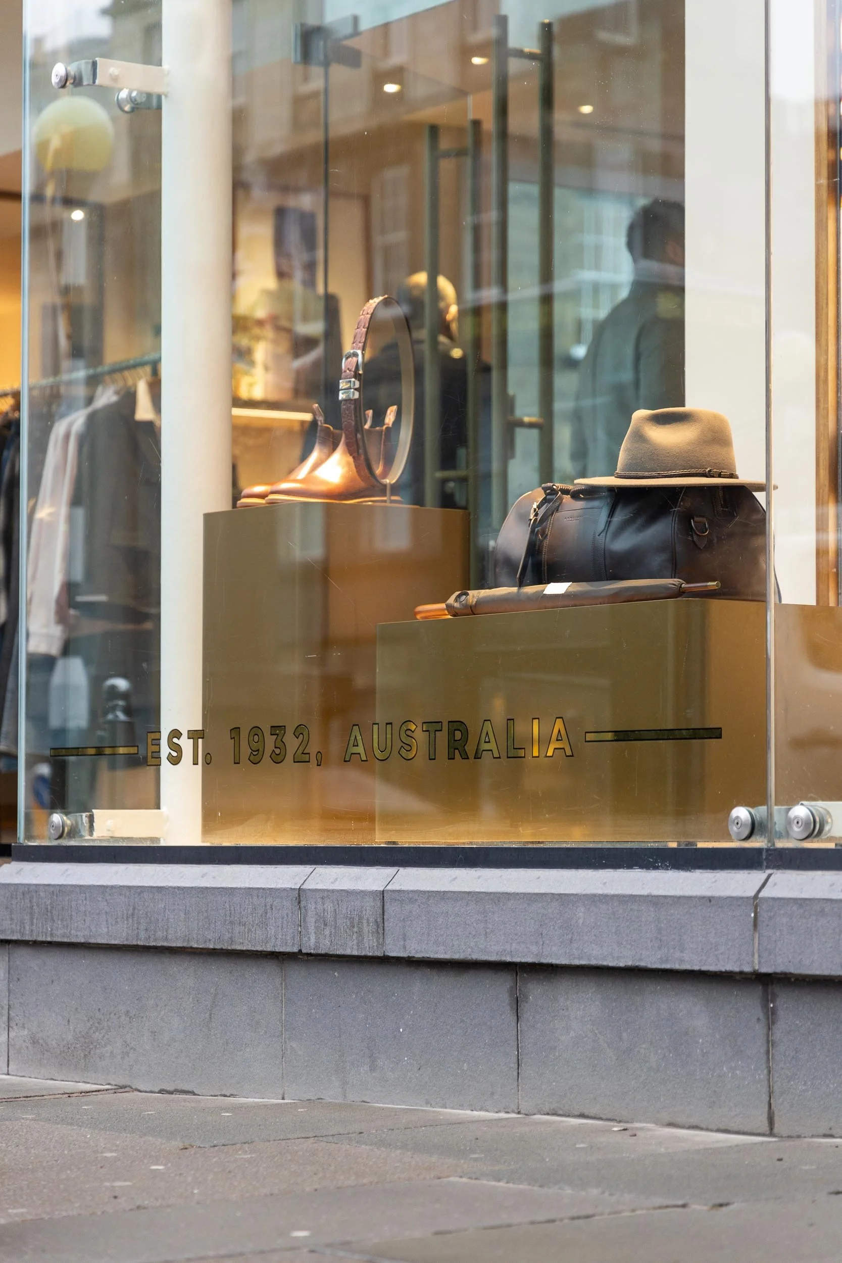 Store display window showcasing men's accessories and clothing, including a belt, boots, a hat, a bag, and a walking stick. The sign indicates the store was established in 1932 in Australia.