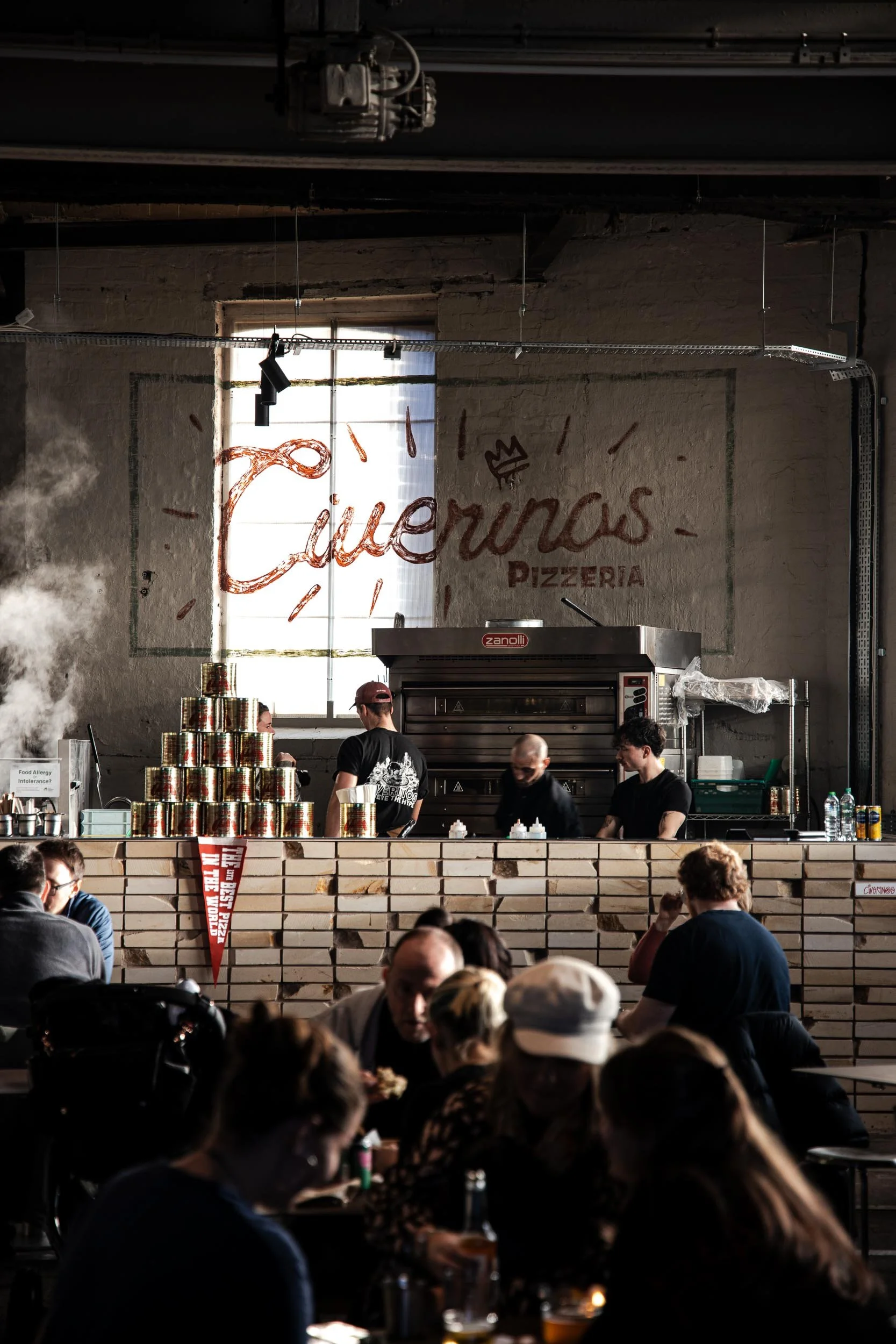 Interior of a pizzeria with a large sign reading 'Cuervos Pizzeria' on the wall behind the kitchen counter. Several staff members are working behind the counter, with stacks of canned tomatoes visible. Customers are seated at tables in the foreground, enjoying their meals.