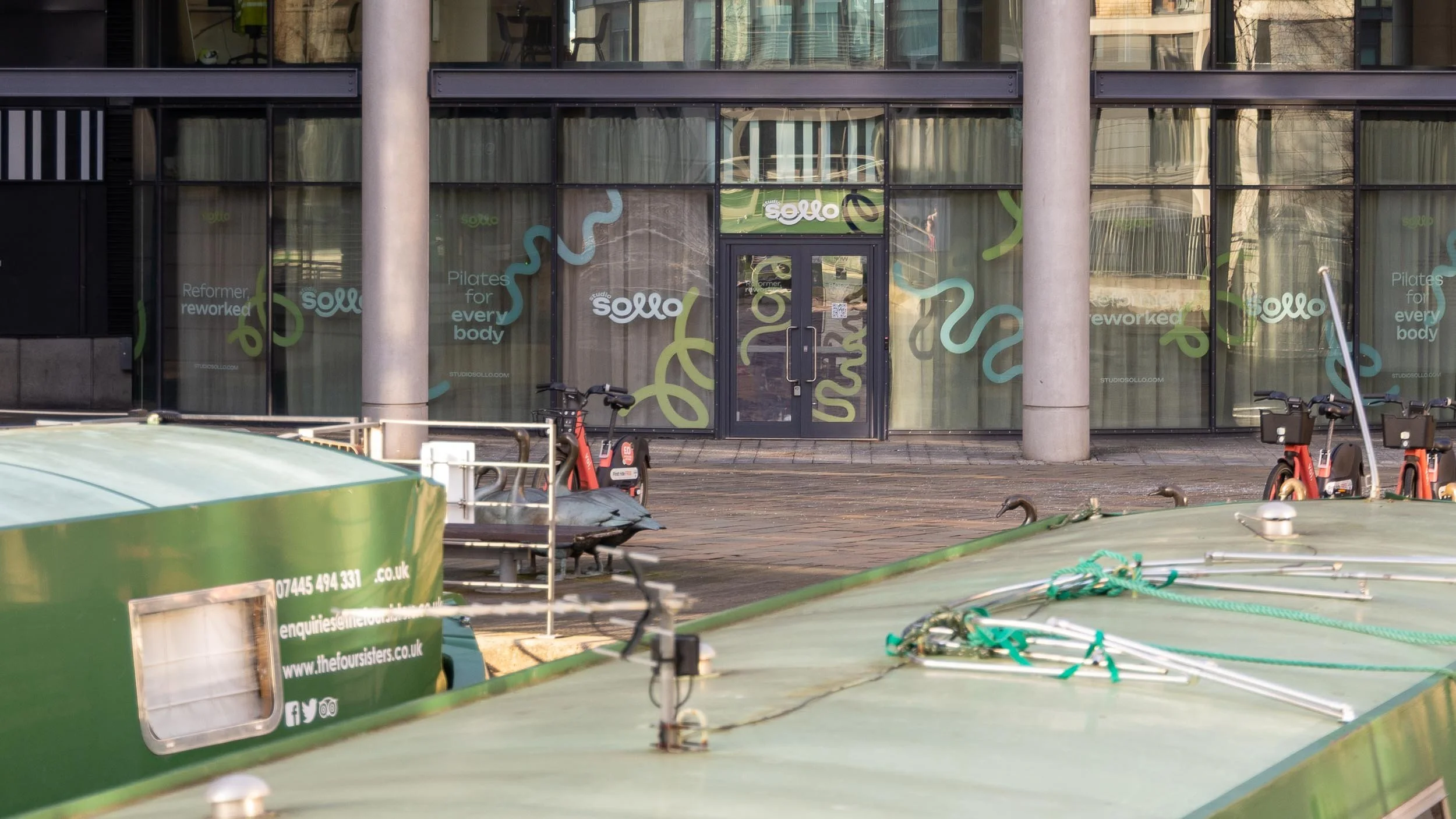 Exterior of a fitness studio called 'Studio Sello' with large windows decorated with colorful swirl designs and text promoting Pilates for all body types, located in an urban area with bikes and a boat in the foreground.