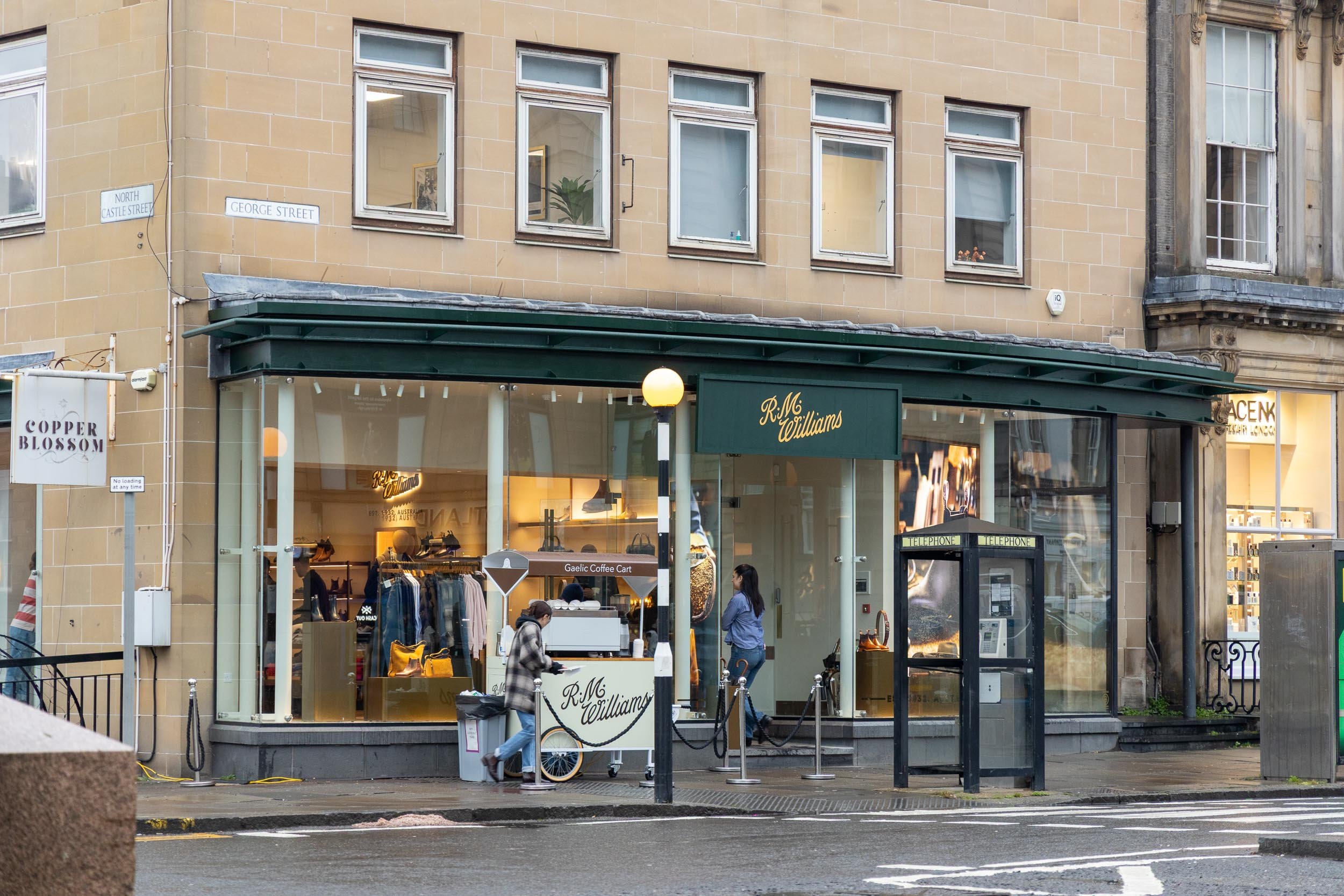 Storefront of R.M. Williams with large glass windows, street signs indicating North Castle Street and George Street, and people in front including a person with a bicycle and another woman talking near a phone booth.