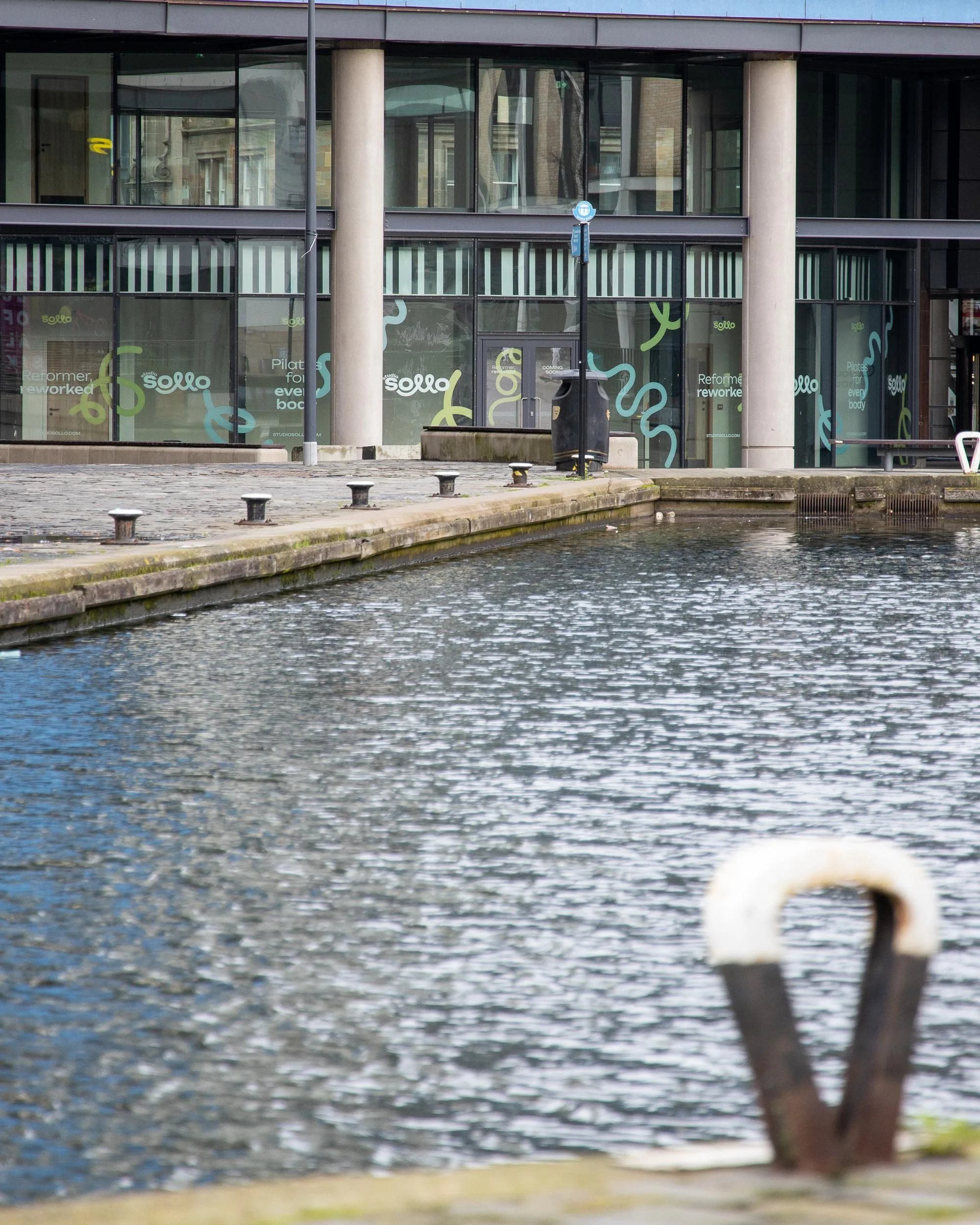 View of a modern glass building reflecting water in the foreground, with graffiti letters on the glass windows reading 'Sollo' and additional text in the background, near a canal or river.