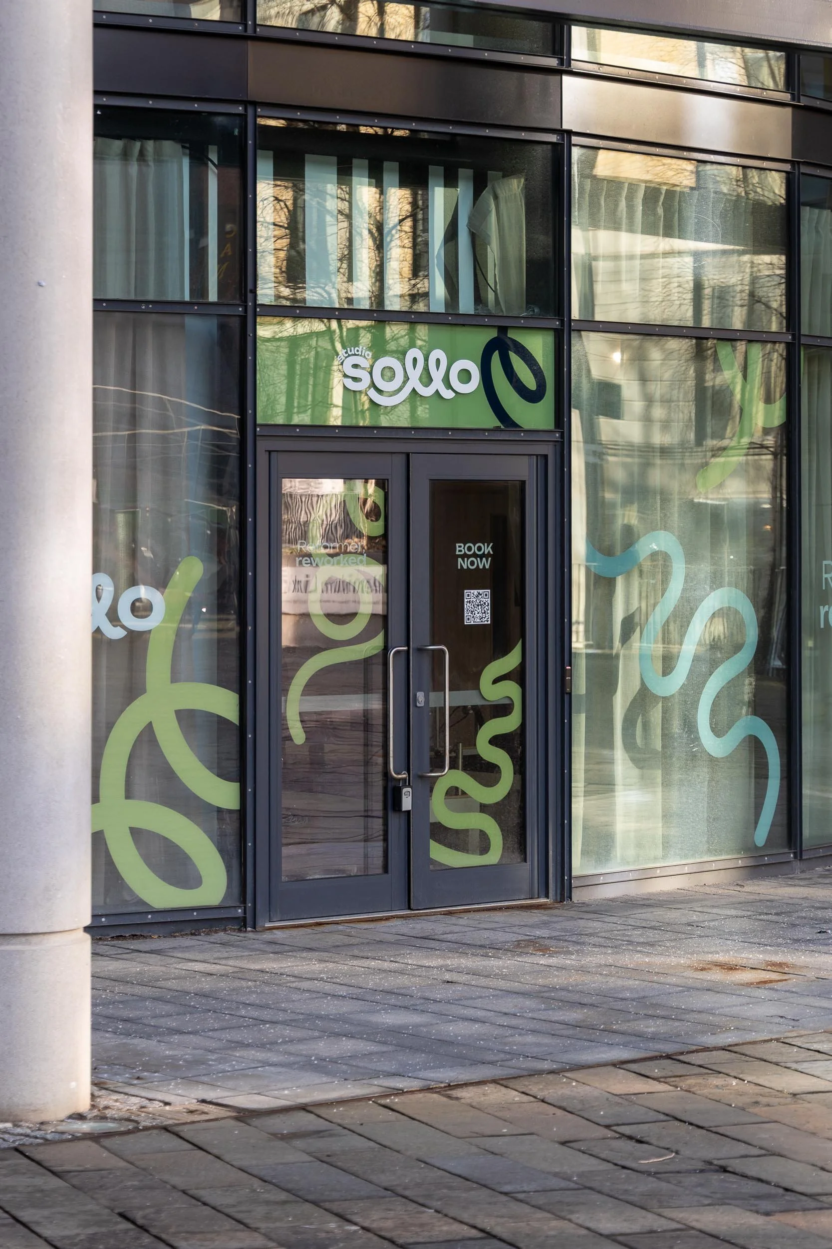 The exterior of a modern building with large glass windows featuring green and black decorative swirls and the sign 'studio sollo.' The entrance door has a sign that says 'BOOK NOW' and a QR code, with some reflections visible in the glass.