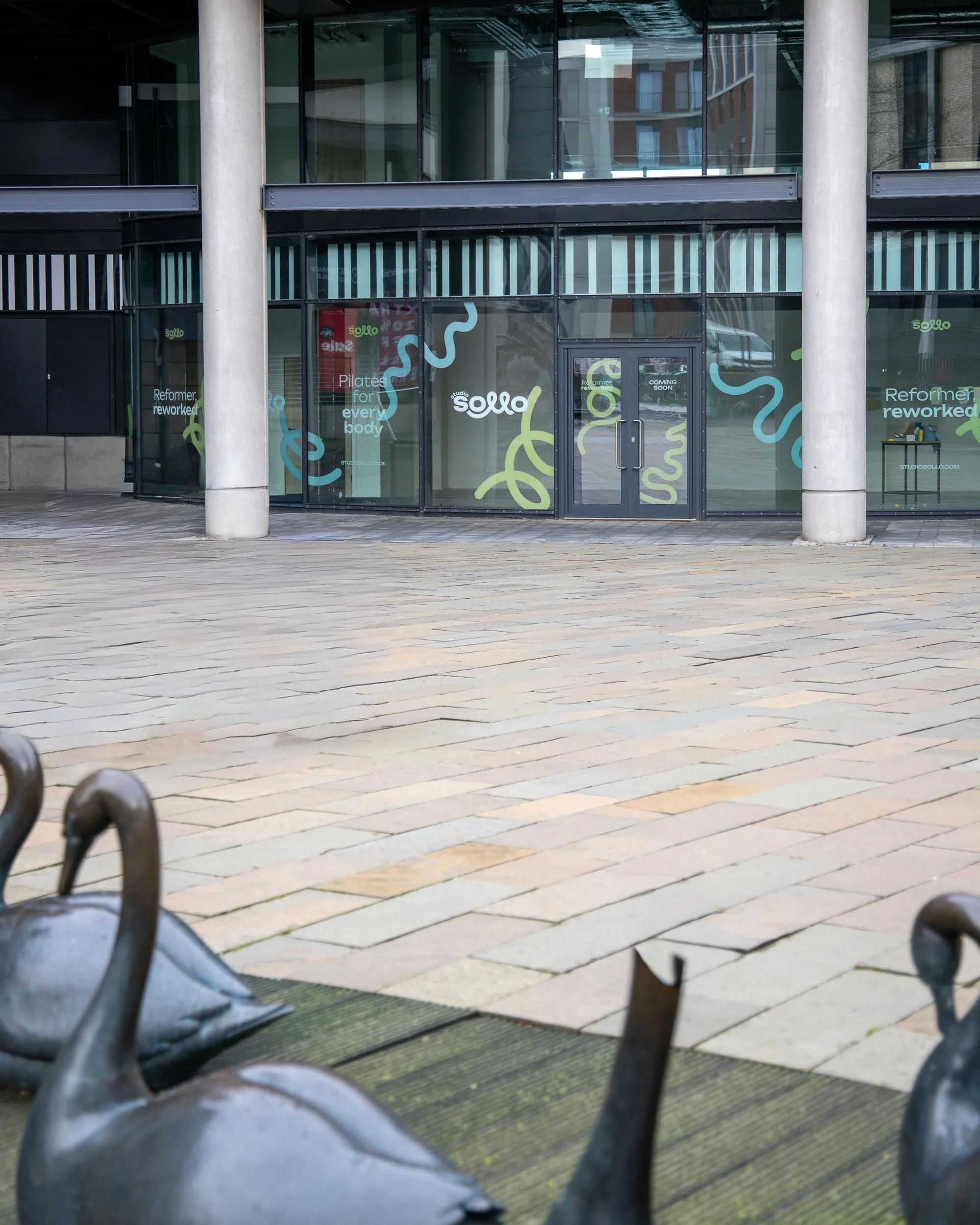 Front of a modern glass building with the logo 'sollo' and colorful abstract designs on the windows, with outdoor metal swan sculptures in the foreground on a paved area.