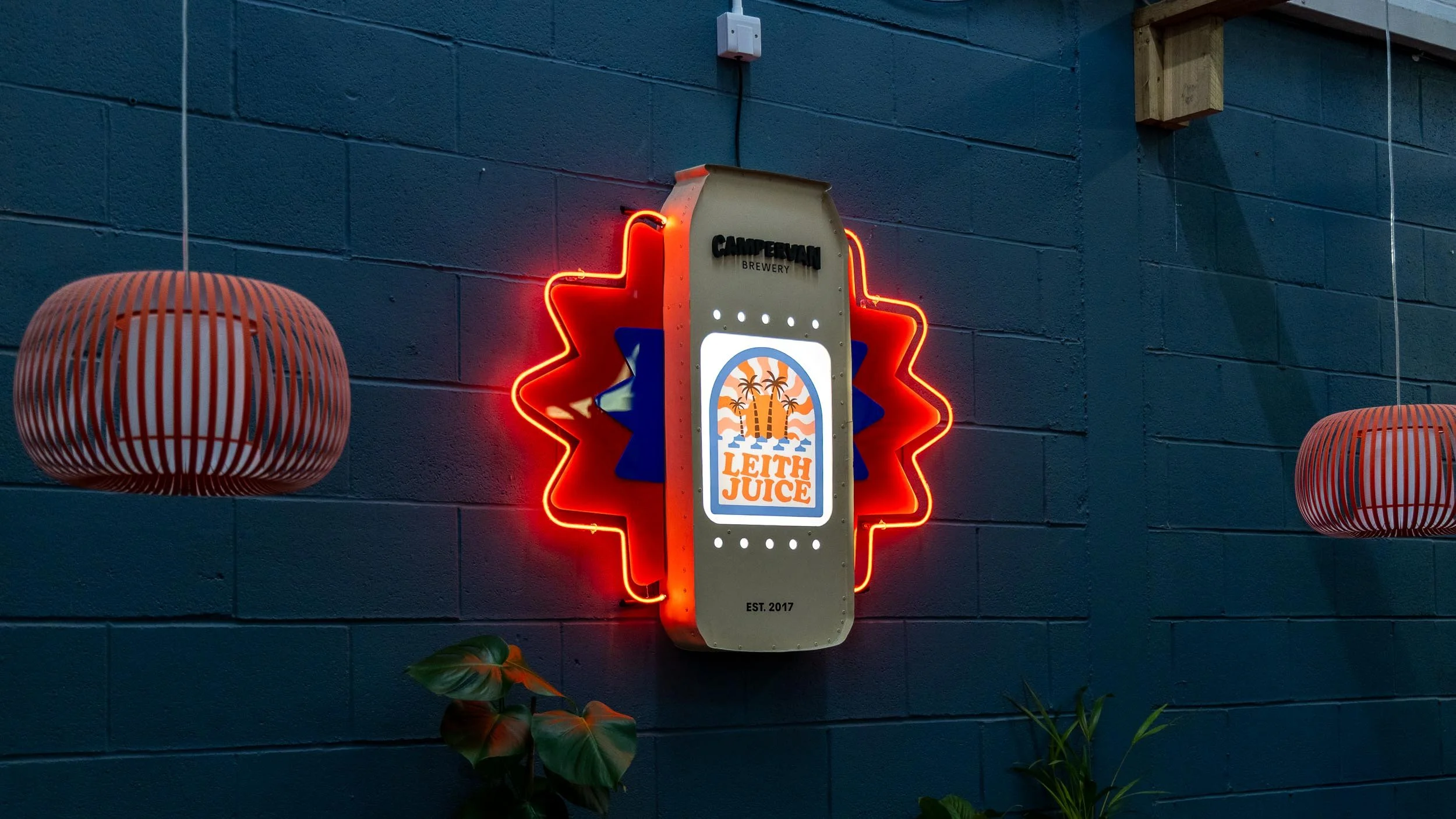 Neon sign shaped like a soda can, with the words 'Campershan Brewery' and 'Leith Juice' on it, mounted on a blue brick wall. Two hanging lanterns are on either side of the sign, and some green plants are at the bottom.