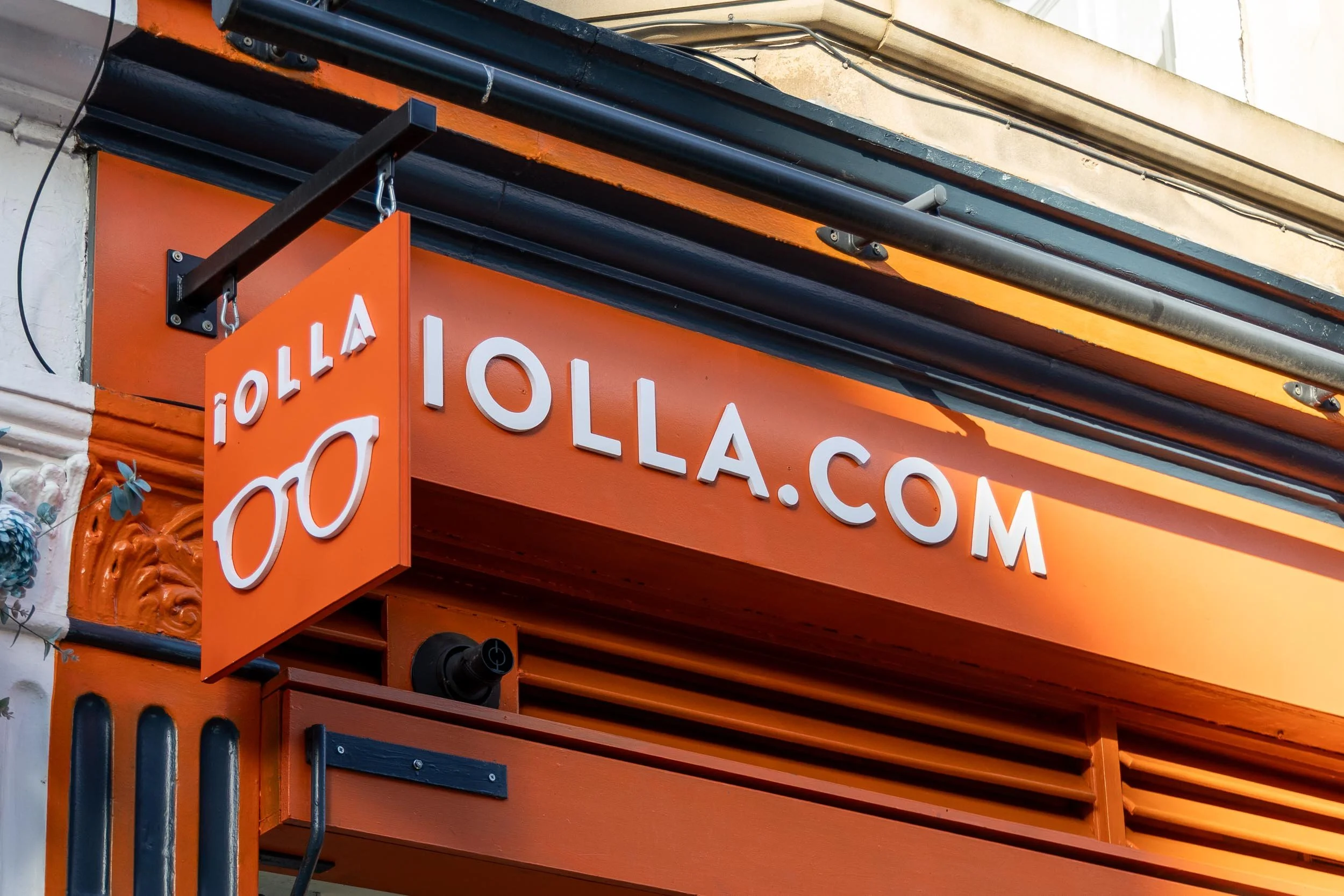 Close-up of an orange exterior sign with white letters displaying 'OLLA.COM' and a small hanging sign with a pair of glasses and the text 'OLLA' in white.