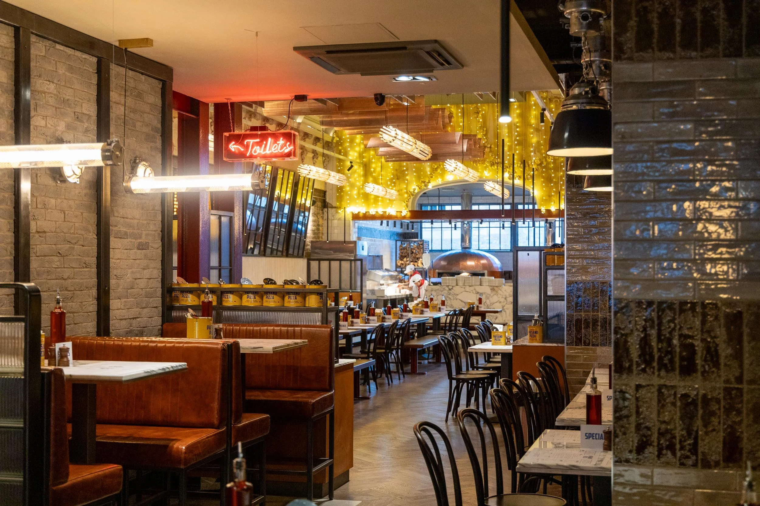 Empty restaurant interior with booths and tables, decorative string lights, a sign pointing to the toilets, and a pizza oven in the background.
