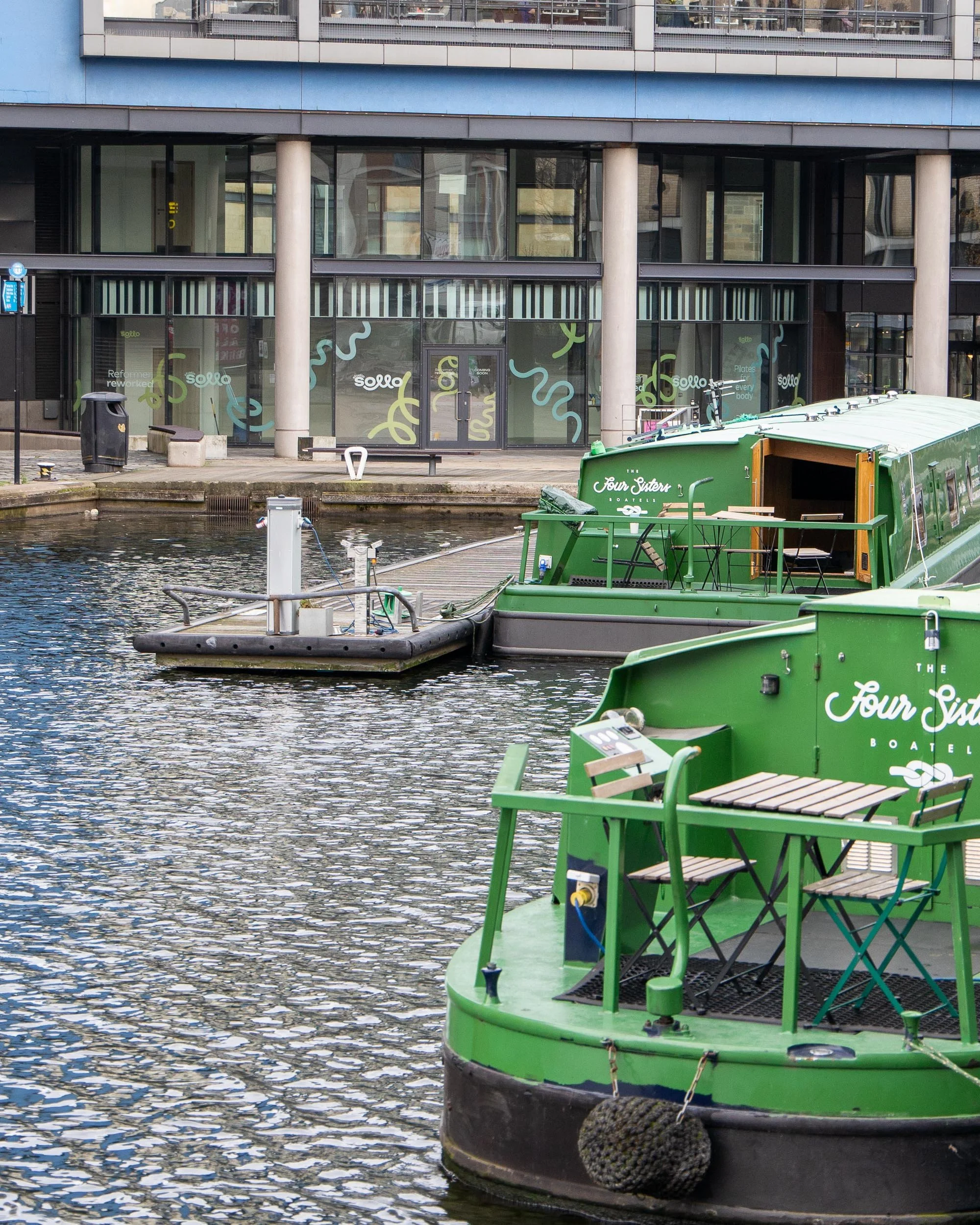 Green boats docked along a canal with a modern building in the background, featuring large windows and colorful signage.