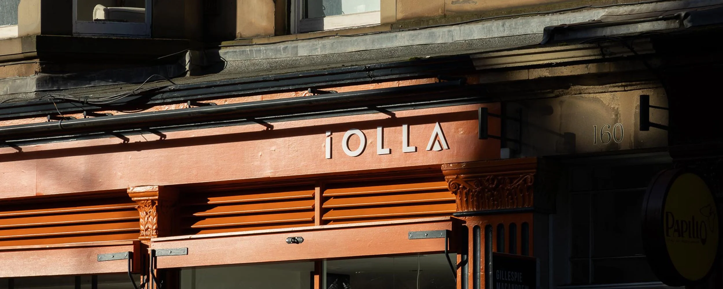 Close-up of a cafe or bar storefront with an orange facade, white lettering spelling "IOU" and decorative architectural details, street number 160, and part of a sign for "Capulo" visible on the right.