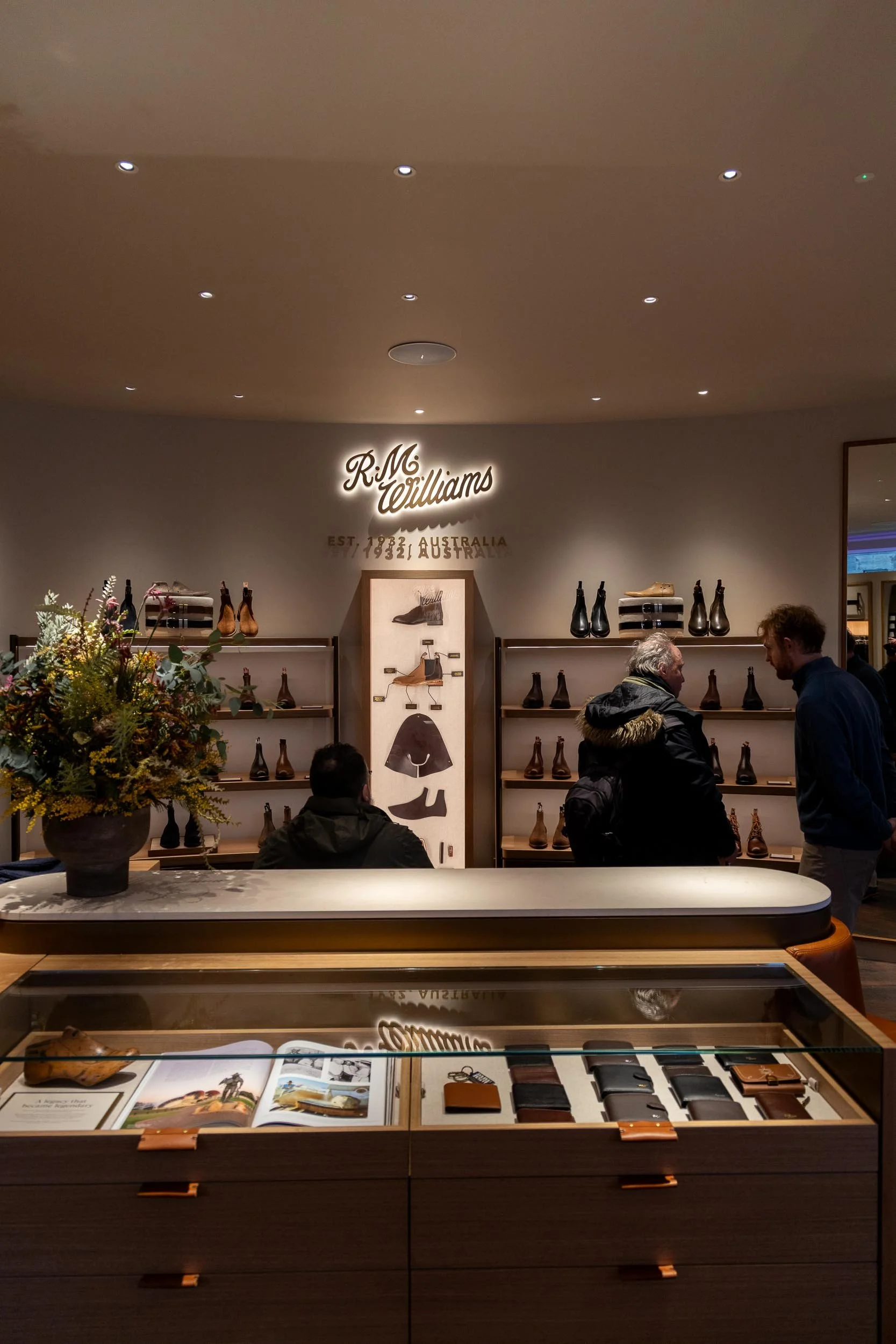 Interior view of a shoe store, RM Williams, with shelves displaying boots, a table with wallets and accessories, and customers shopping. Centred to the product display the back-lit sign mounted on the wall saying " R.M.Williams"