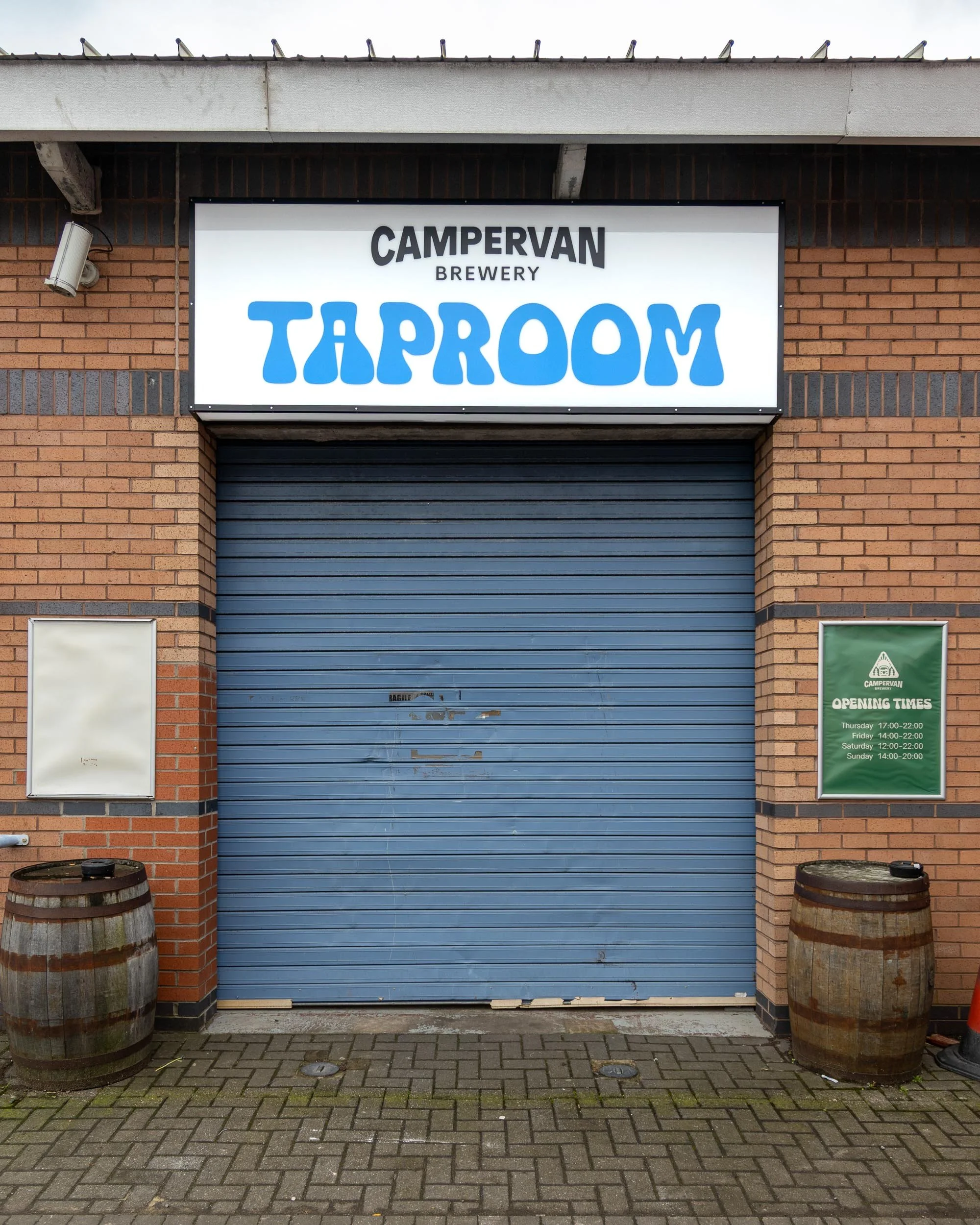 Closed blue rolling door with a sign above it that reads 'CAMPERVAN BREWERY TAPROOM' in large blue and black letters, and a green sign on the right displaying opening times for Thursday through Sunday.