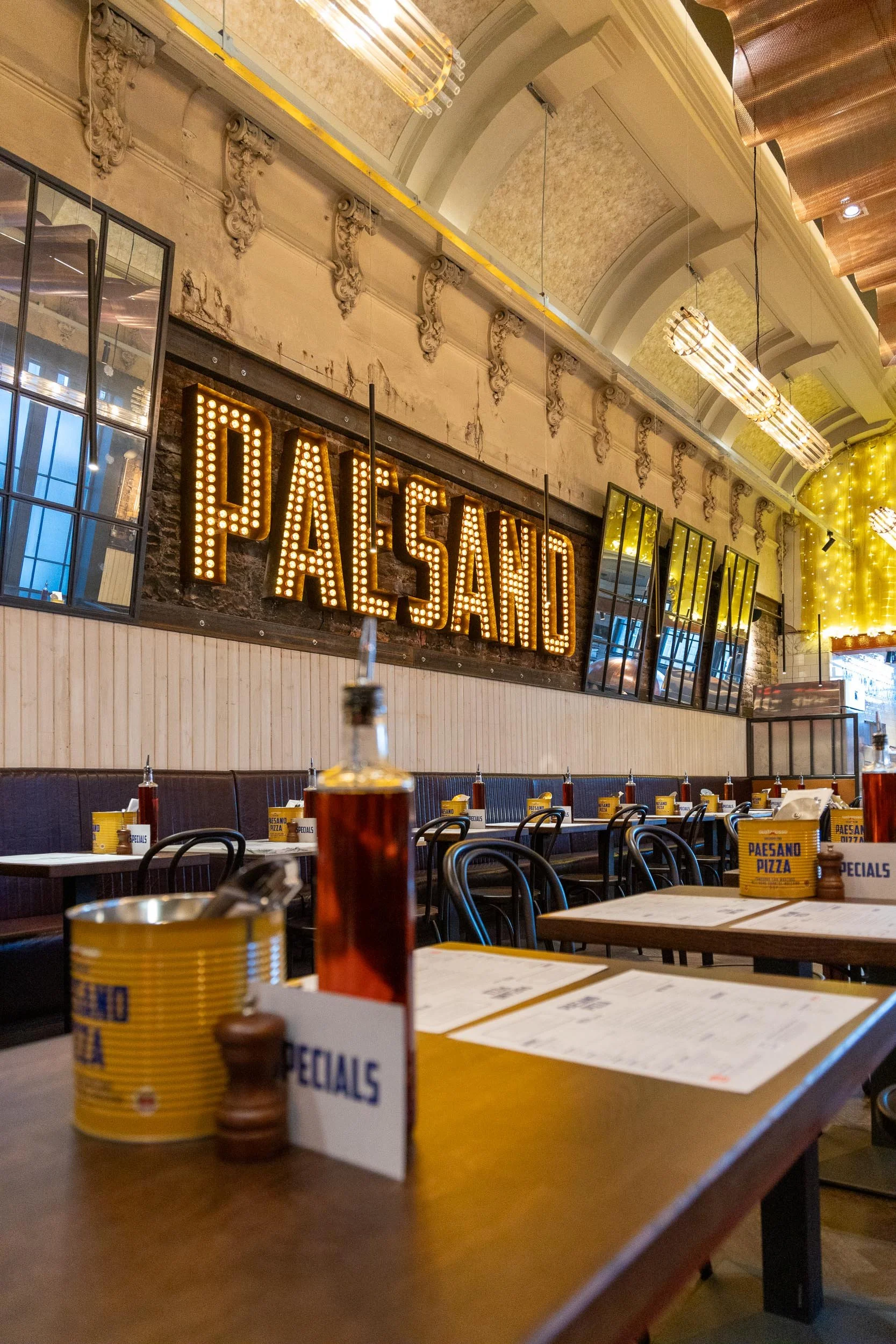 Interior of a restaurant called Paesano, with a large illuminated sign spelling 'PAESANO' on the wall, tables set with menus, condiments, and bottles of hot sauce, and decorative lighting fixtures hanging from the ceiling.