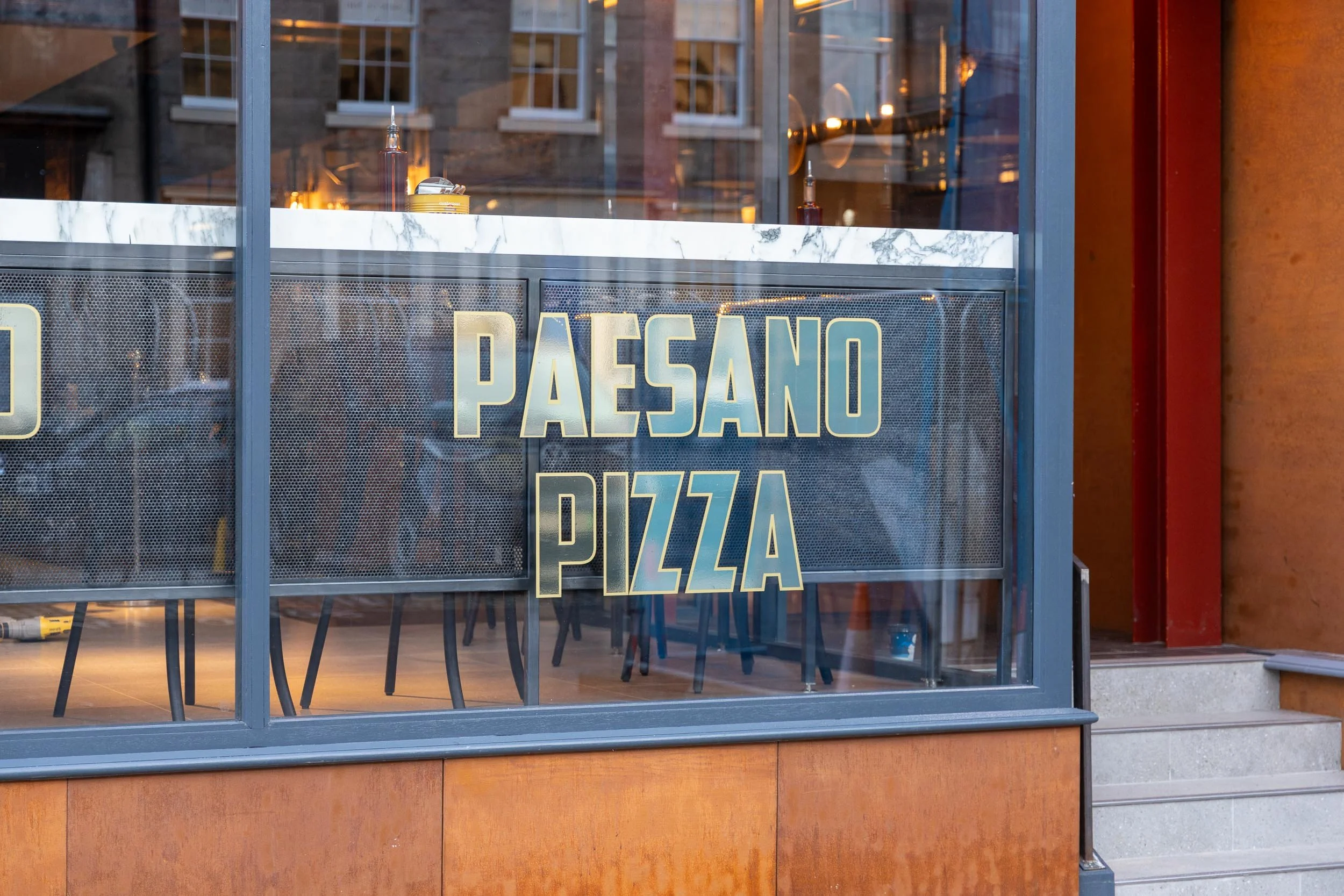 Storefront window with a sign that reads 'PAESANO PIZZA', showing interior tables and chairs through the glass.