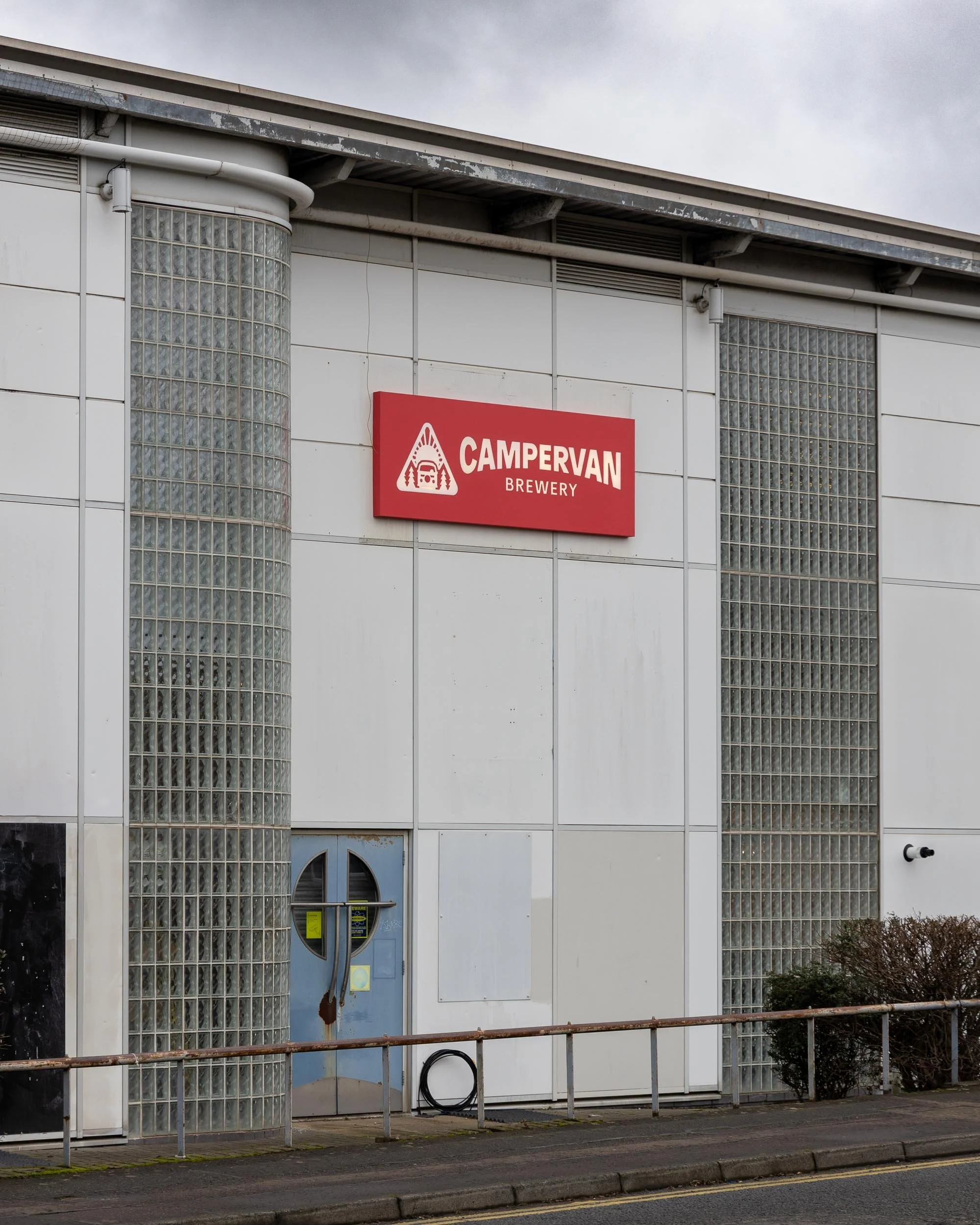 Exterior view of a building with white walls, glass block windows, and a sign for Campervan Brewery. The sign is red with white lettering and logo, mounted on the wall above a blue circular door with a metal handle.