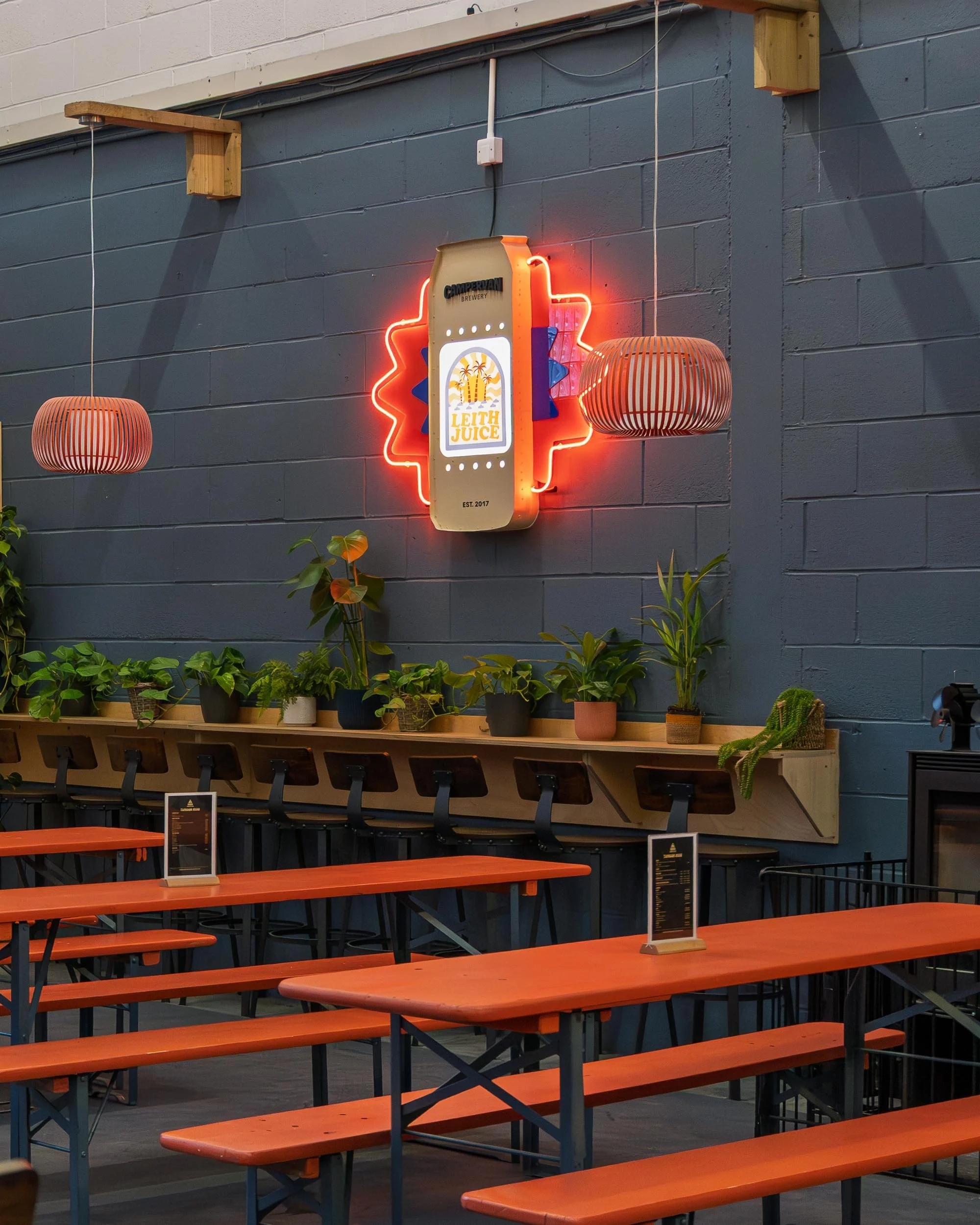 Indoor seating area with orange picnic tables and black metal chairs, decorated with potted plants on a wooden shelf against a dark gray wall. A neon sign shaped like a juice bottle with the brand name 'Leith Juice' and bright lights is mounted on th