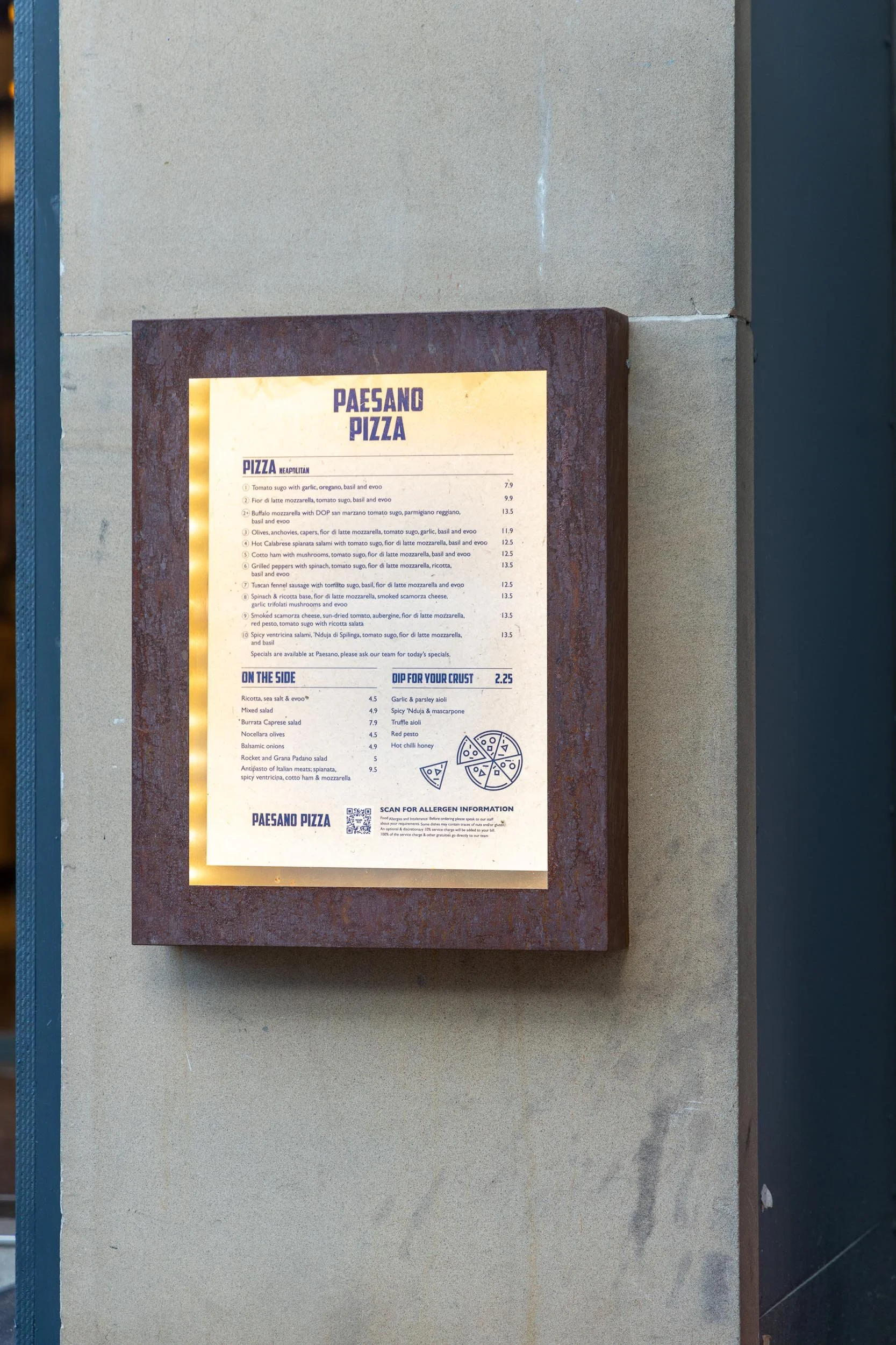 A restaurant menu board displaying various pizza options, side dishes, and dipping sauces, mounted on a textured wall.