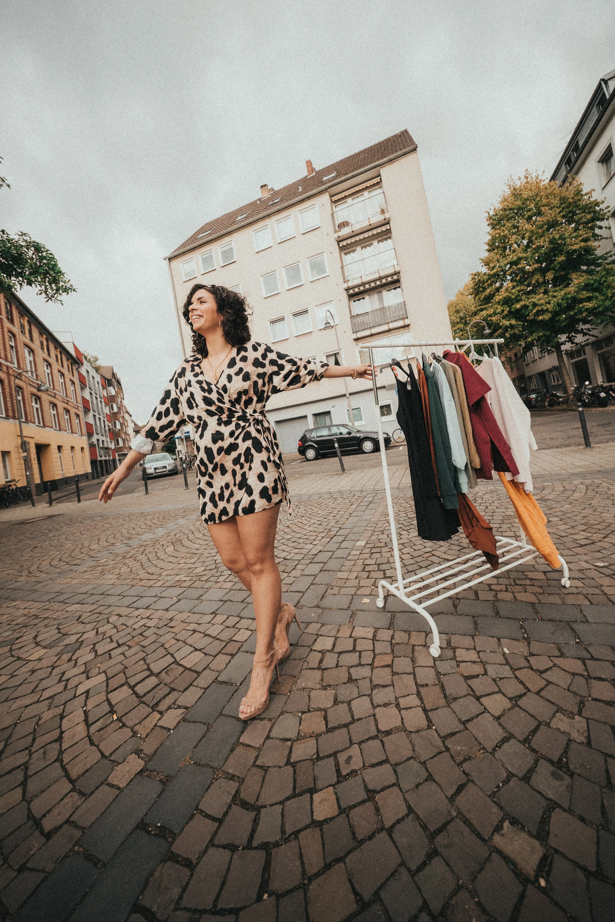 A woman in a leopard print dress and high heels is smiling and walking on cobblestone street with a clothing rack on wheels filled with various pieces of clothing.