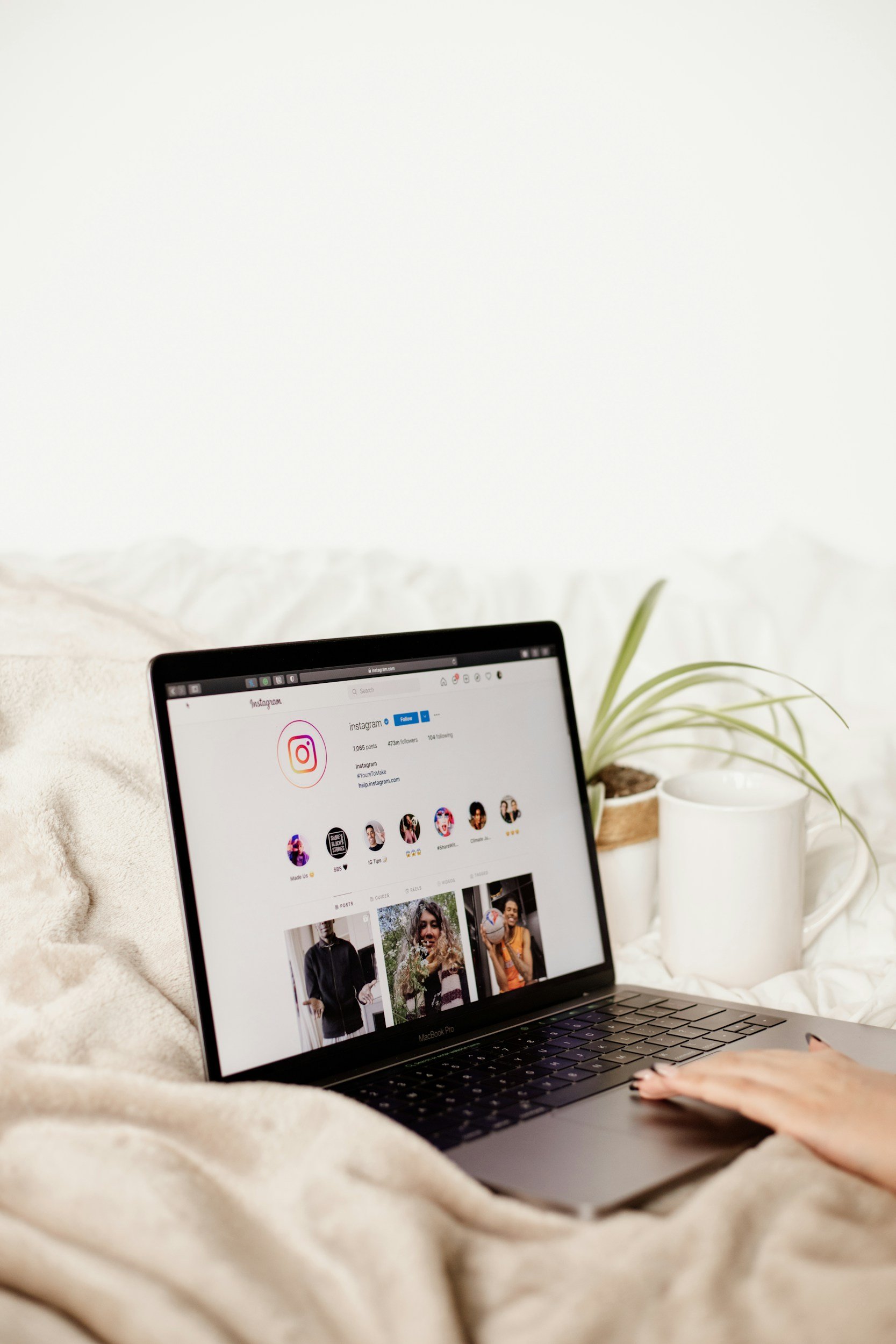 Laptop displaying Instagram profile on a bed with white blankets, surrounded by a plant in a white pot and a white mug.
