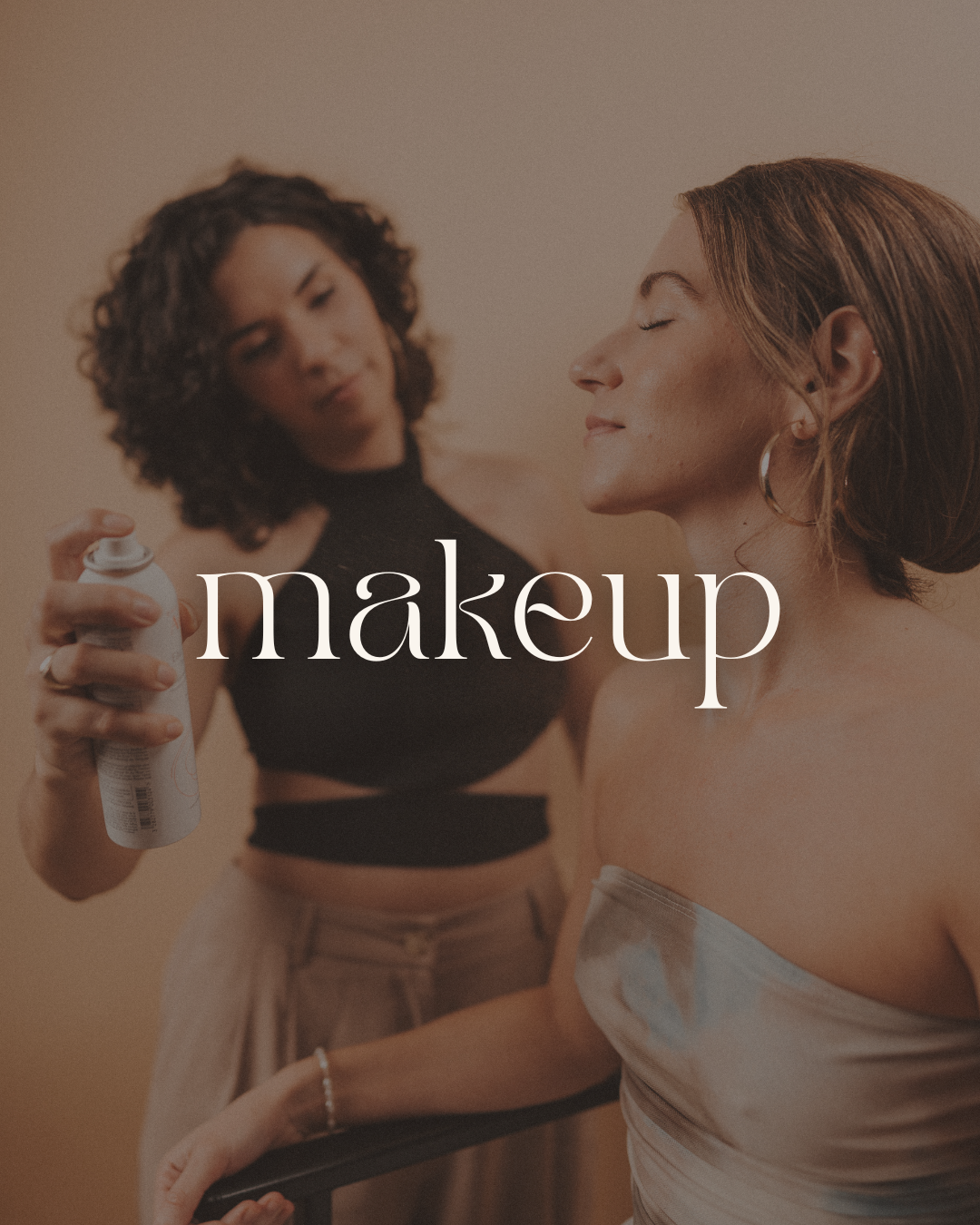 A makeup artist applying spray makeup on a woman with closed eyes and a content expression, while holding a spray can. The woman has short hair, large hoop earrings, and wears a strapless top. The background is neutral-colored.