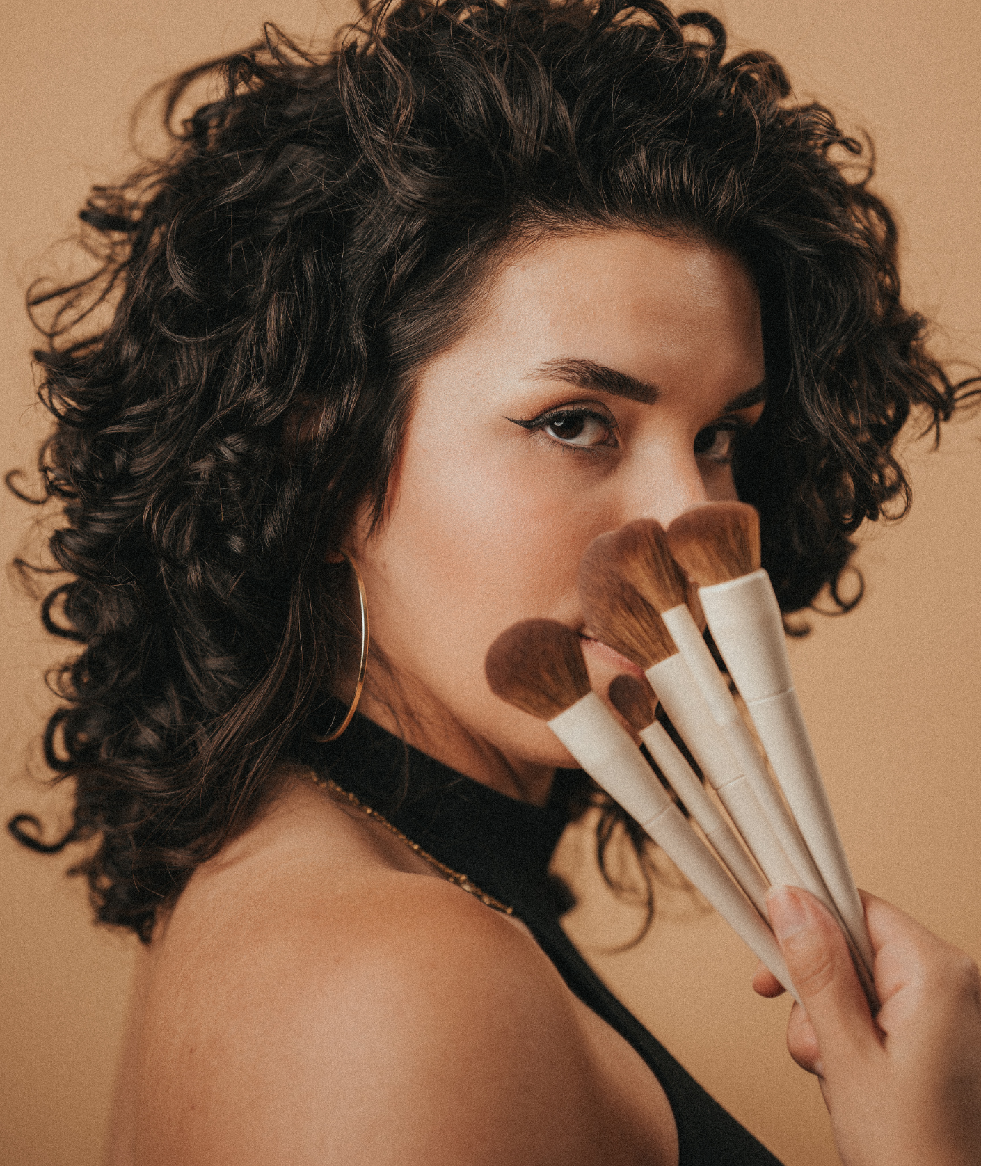 A woman with curly dark hair holds makeup brushes in front of her face, partially covering her mouth and nose, against a neutral background.