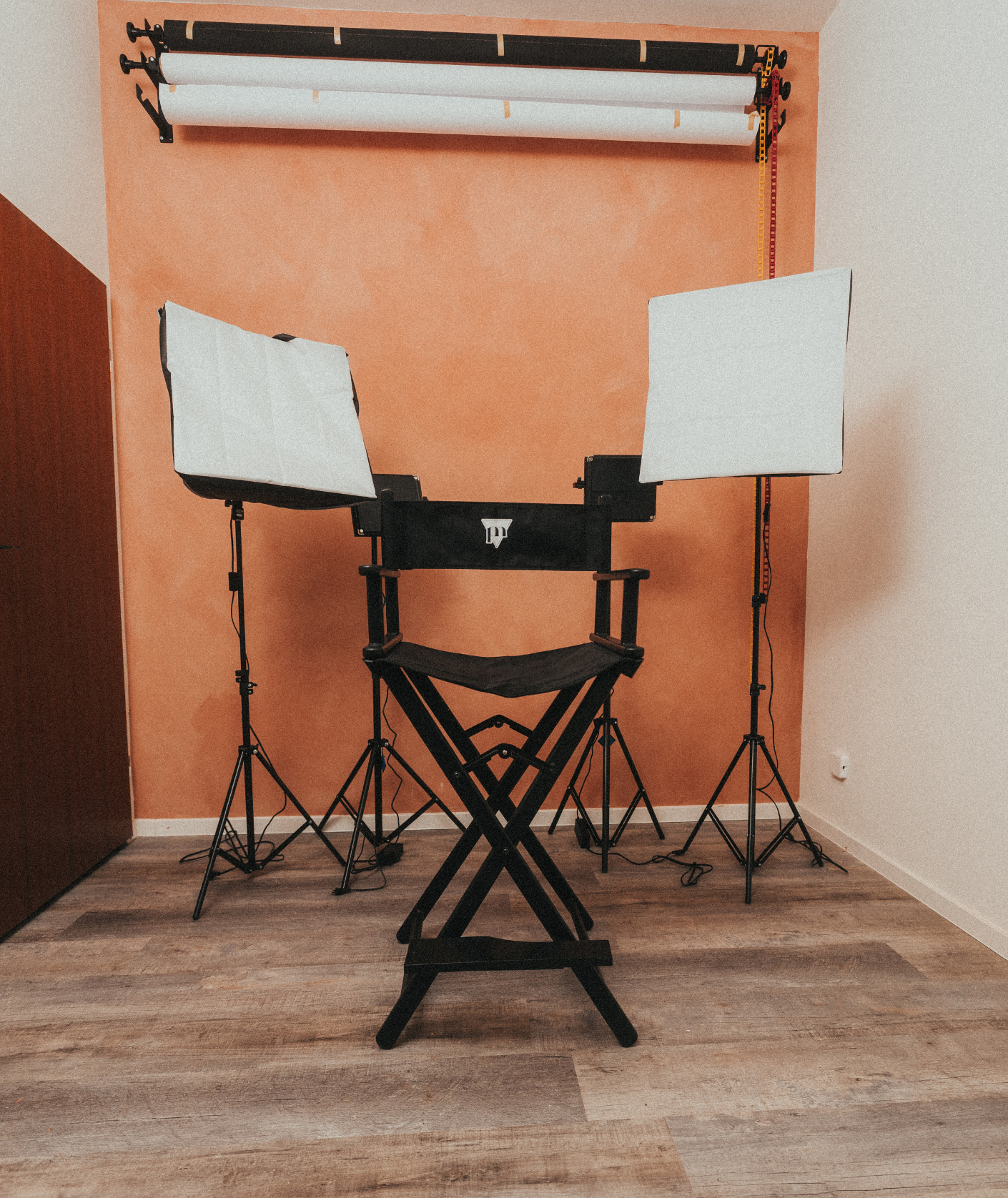Photography setup including a camera on a tripod, two softbox lights, a backdrop with orange walls, and two large rolls of photography paper, in a room with wooden flooring.
