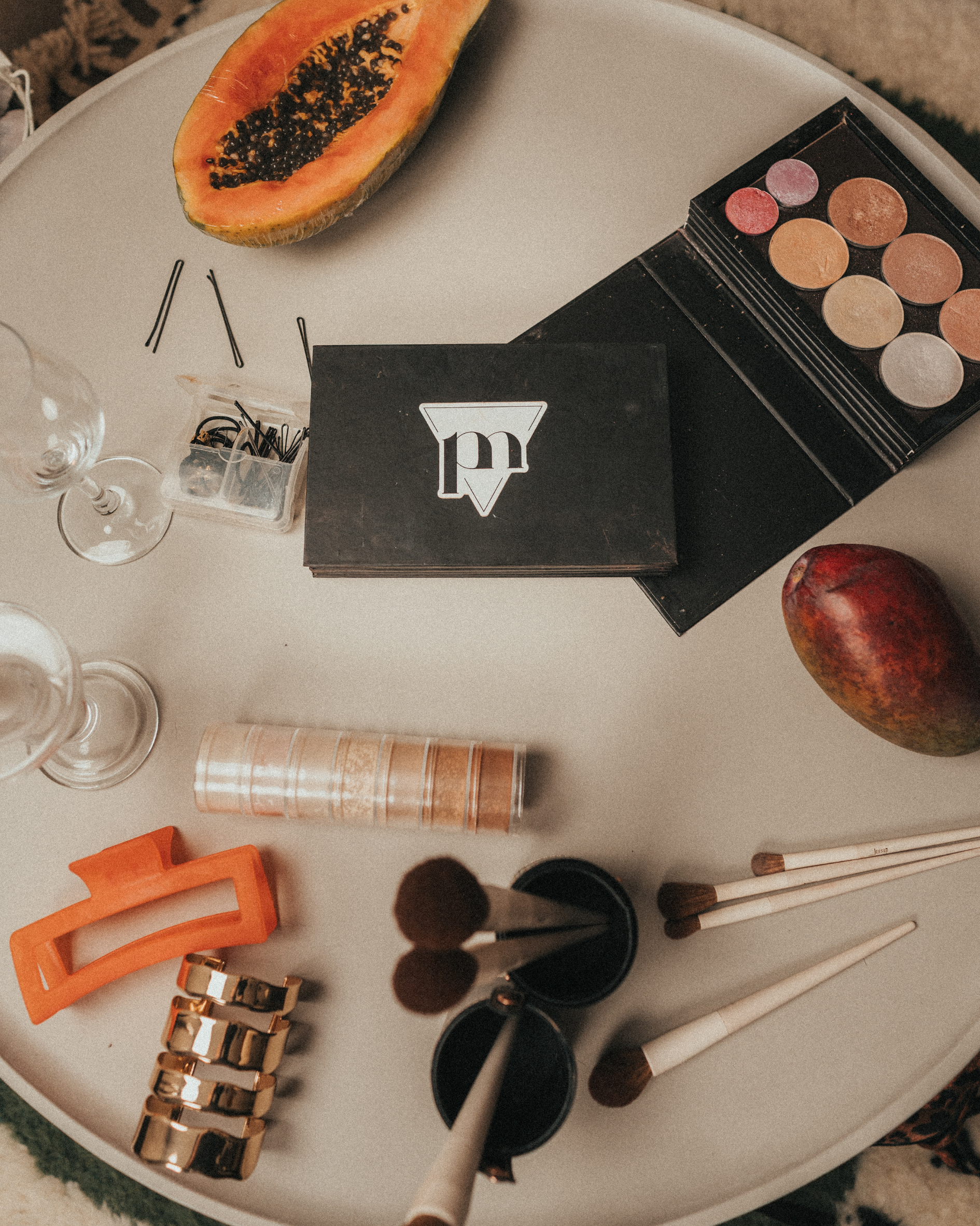Flat lay of various makeup and beauty items including a half cantaloupe, eyeshadow palette, hair clips, glasses, and makeup brushes on a white tray.