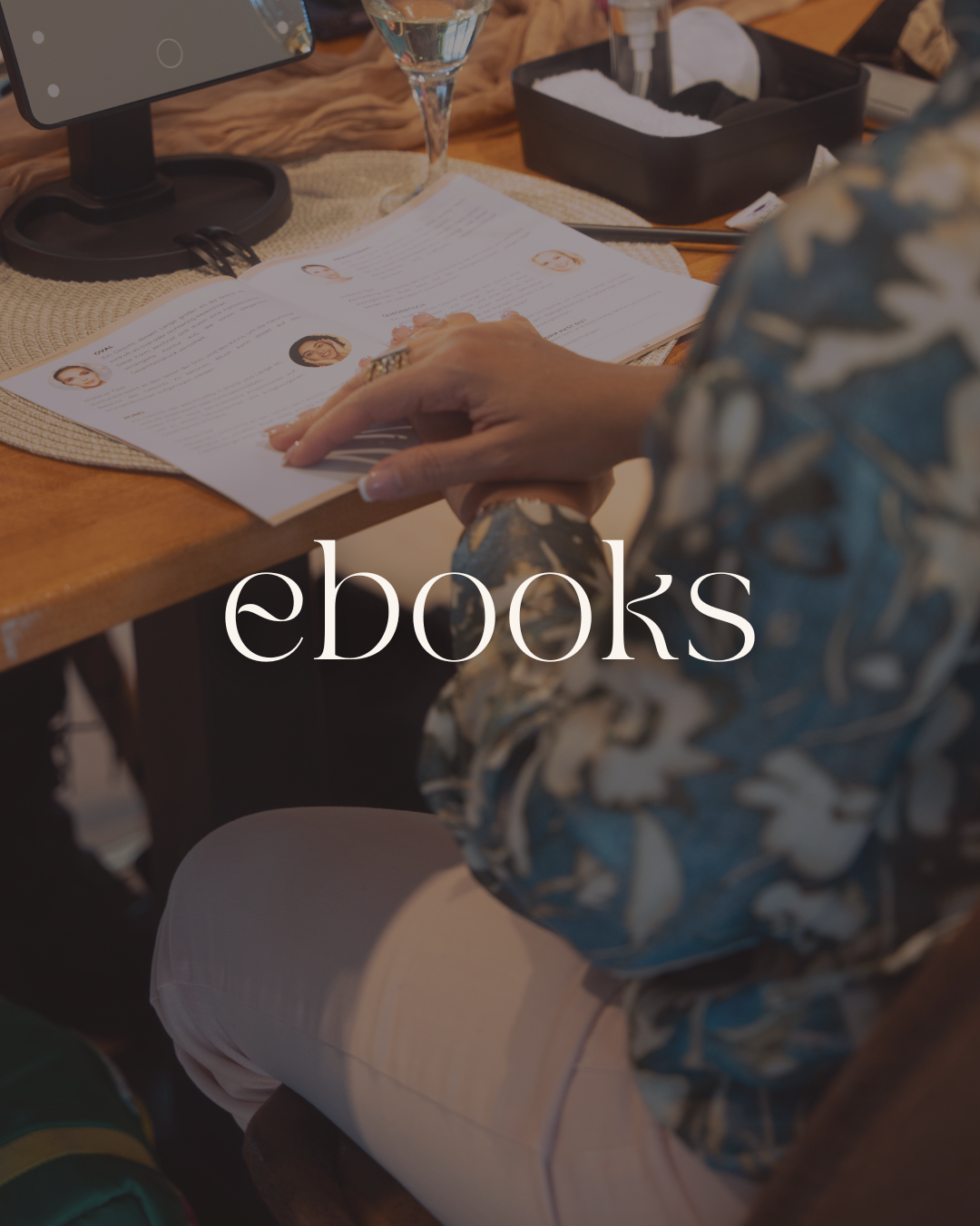 A person looking at an open eBook with author and character photos on a wooden table, surrounded by a glass of water, a monitor, and a black container with items inside.