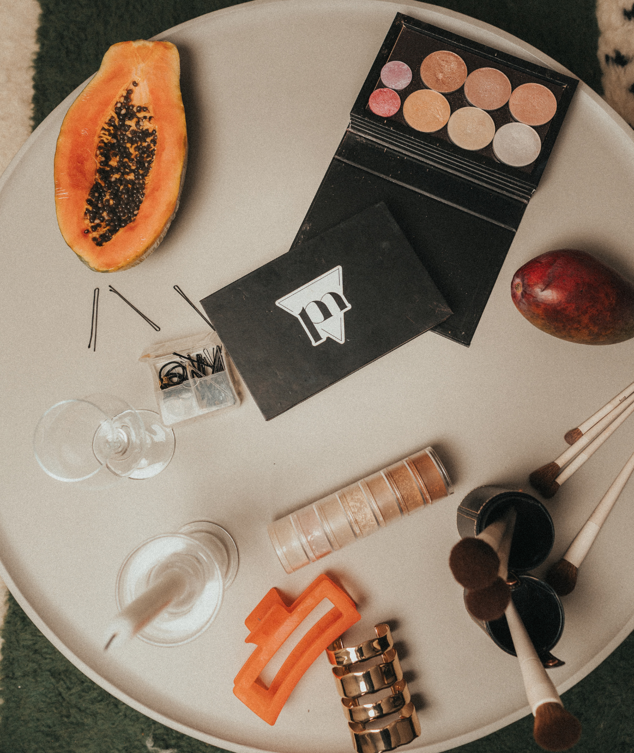 A round table with a halved papaya, a mango, makeup palette, drink glasses, hair clips, makeup brushes, and various hair accessories.
